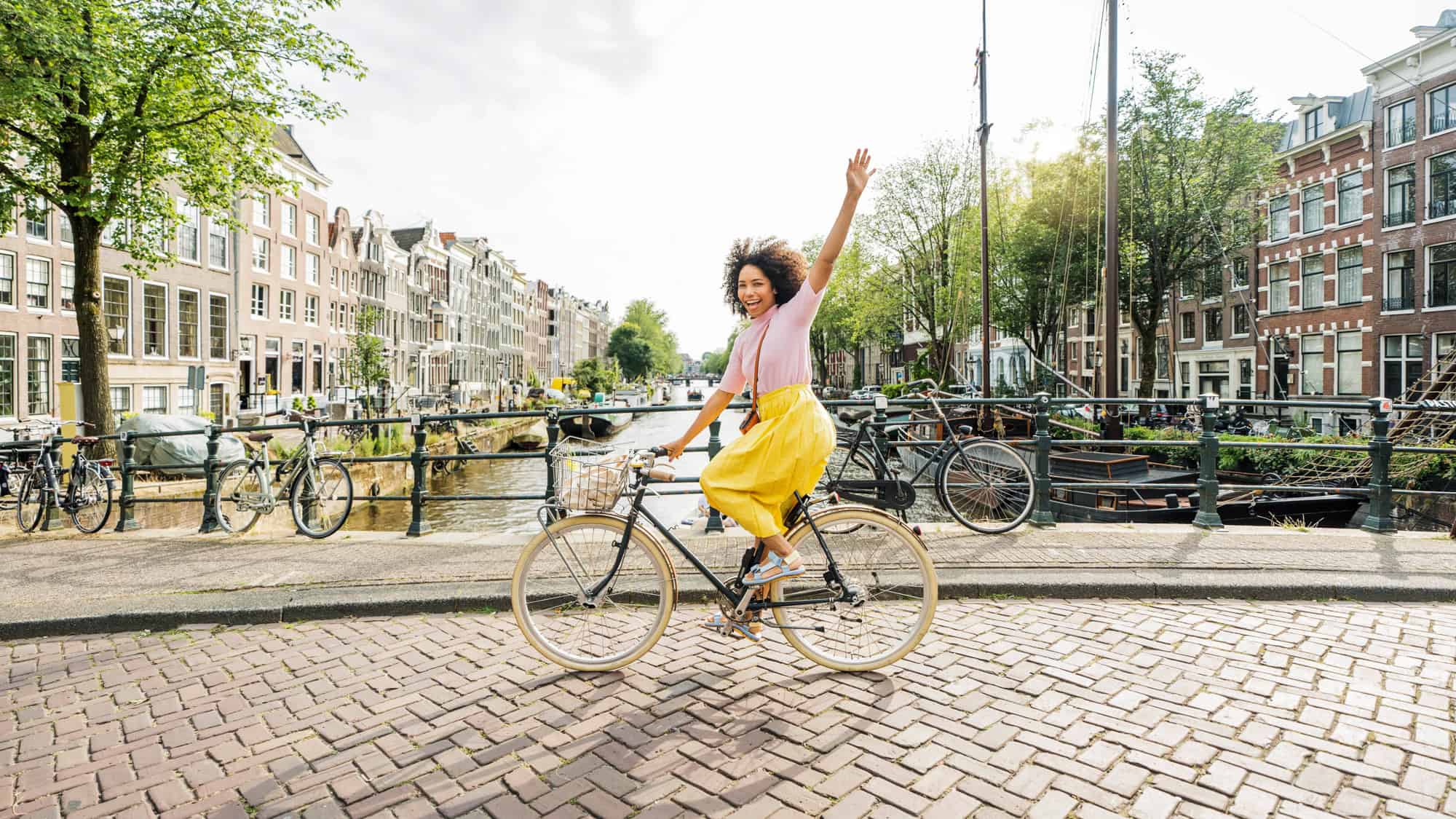 A smiling woman in a yellow skirt rides a bicycle one-handed across a cobblestone bridge over a canal in Amsterdam, surrounded by classic Dutch buildings and parked bikes.