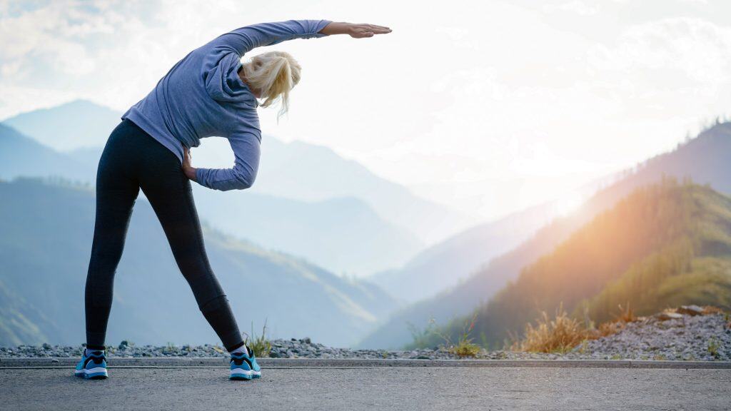 A woman in athletic wear stretches her side on a mountain road with scenic layered hills and a glowing sunrise in the distance.