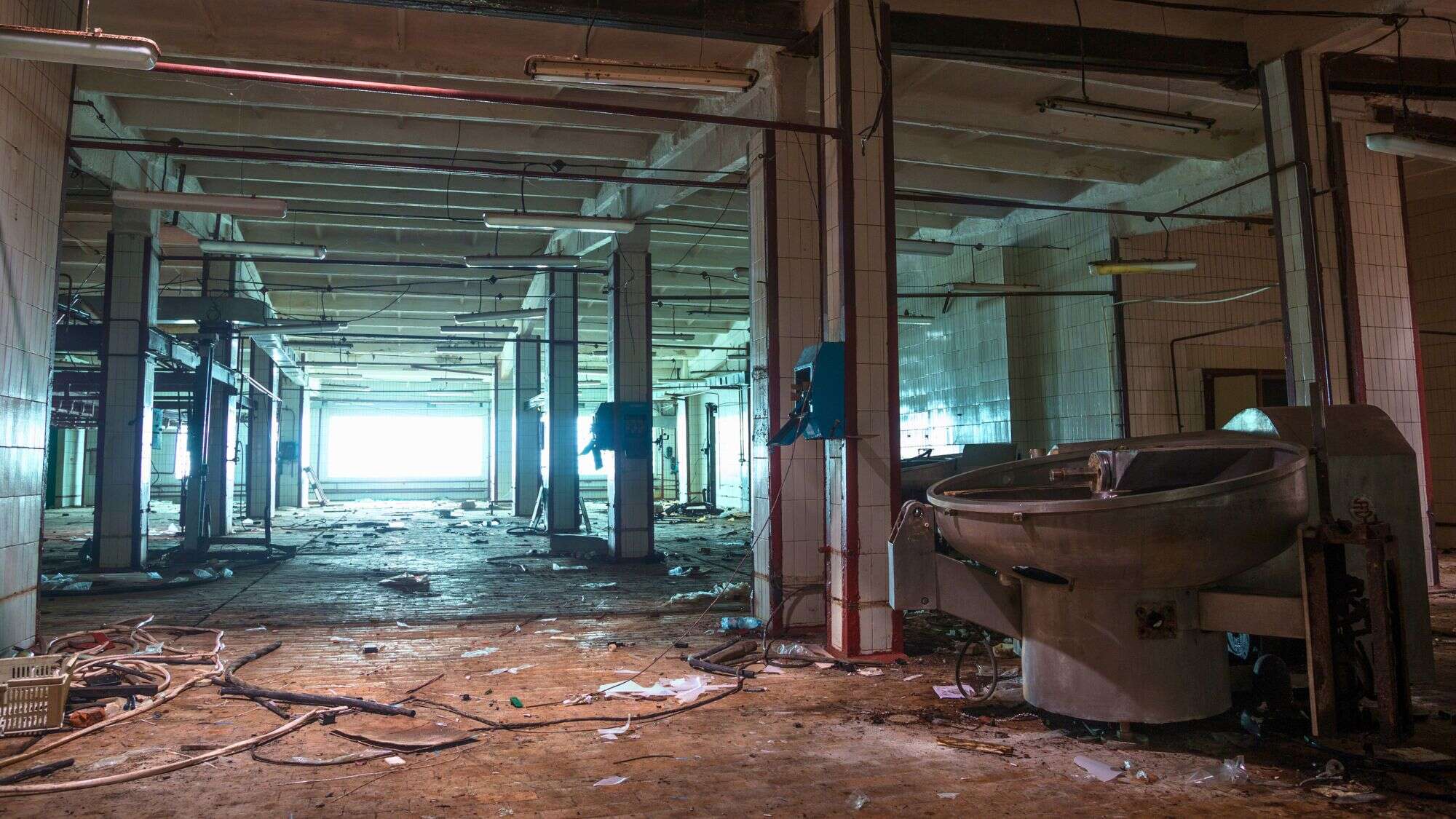 A wide, cluttered view of a large, abandoned industrial kitchen or factory floor, with debris, rusted machinery, and overhead pipes under harsh fluorescent lighting.