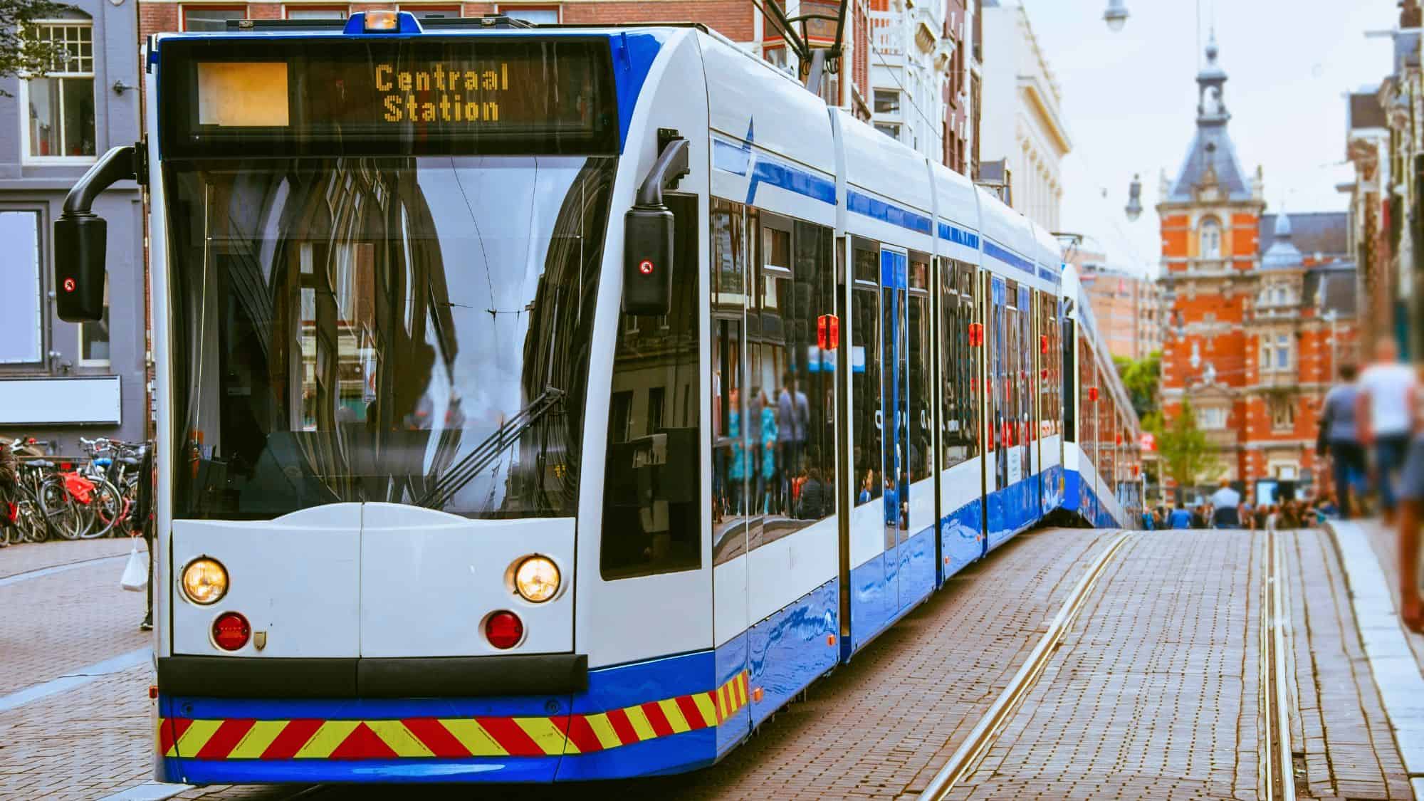 A blue and white tram marked “Centraal Station” travels down a cobblestone street with bicycles and historic buildings nearby.