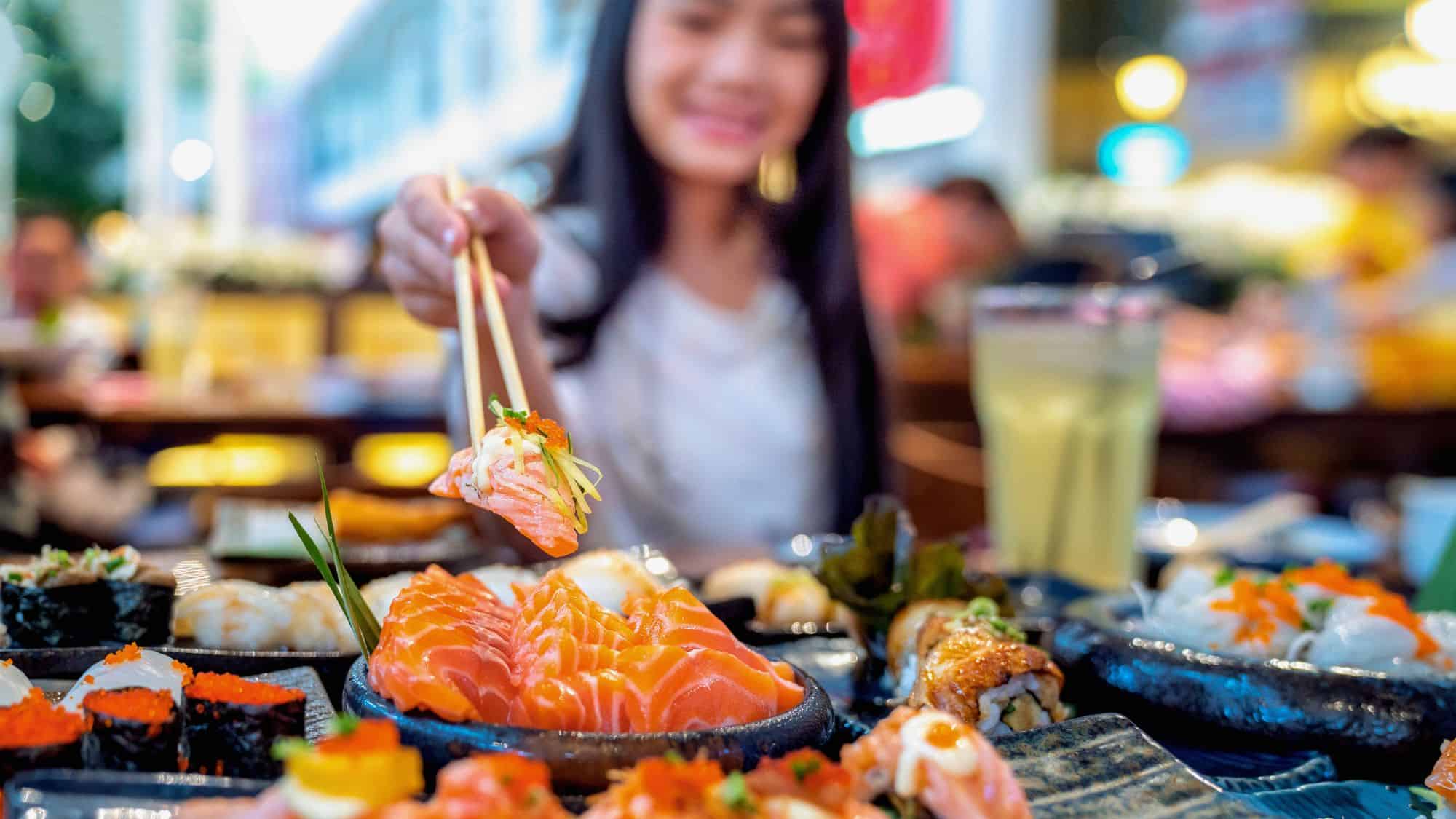A woman uses chopsticks to pick up a piece of salmon sushi, with a table full of assorted sashimi, rolls, and nigiri in front of her.
