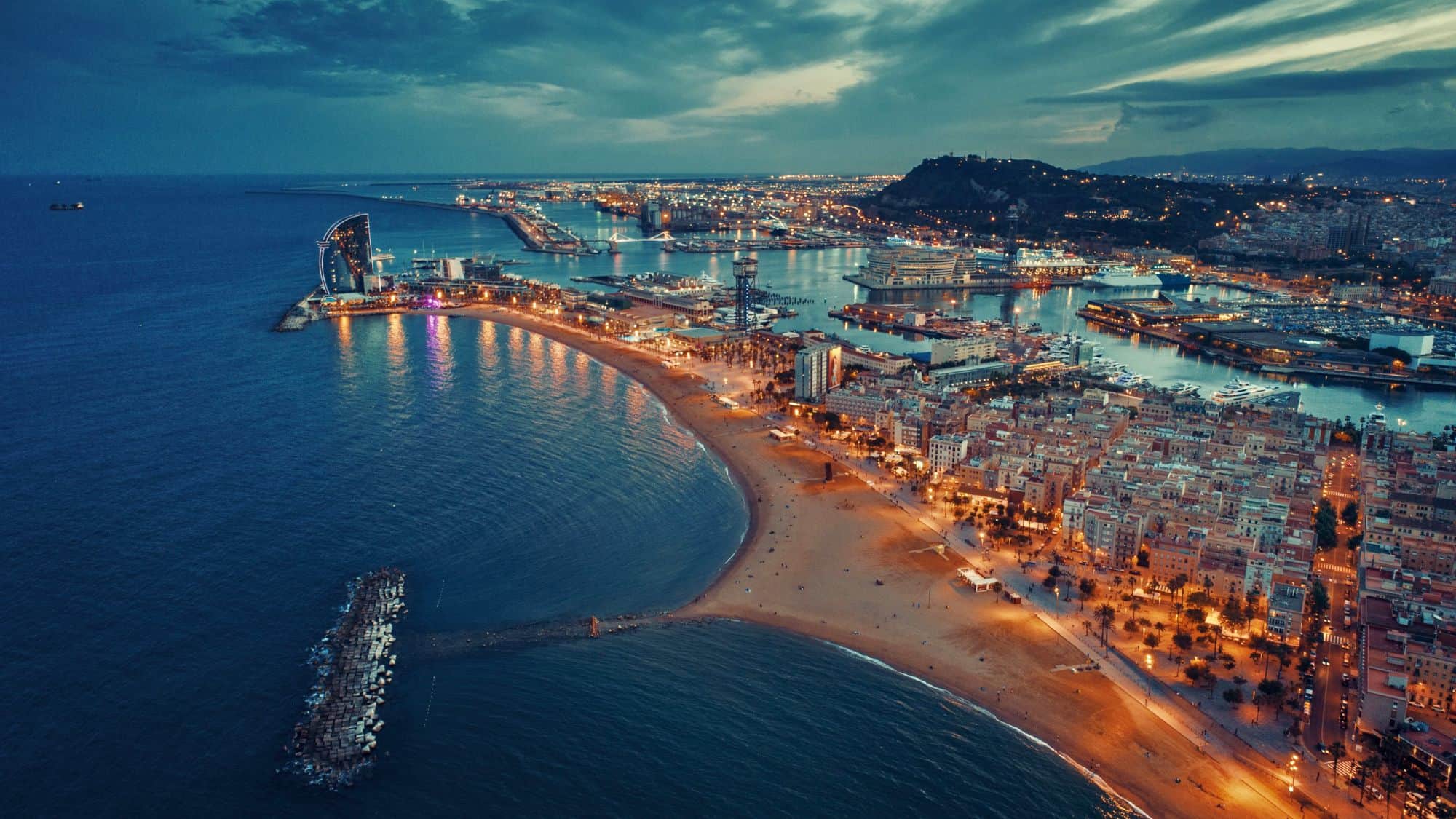 Aerial view of Barcelona at dusk with city lights glowing along the beach and the sea stretching into the horizon.