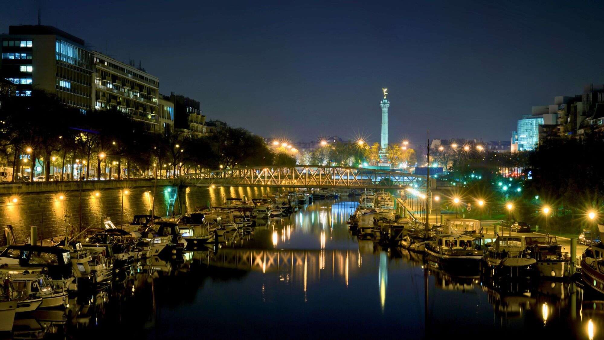 Moored boats line the calm waters of the Port de l'Arsenal in Paris, with the illuminated July Column of Place de la Bastille glowing in the distance under a clear night sky.