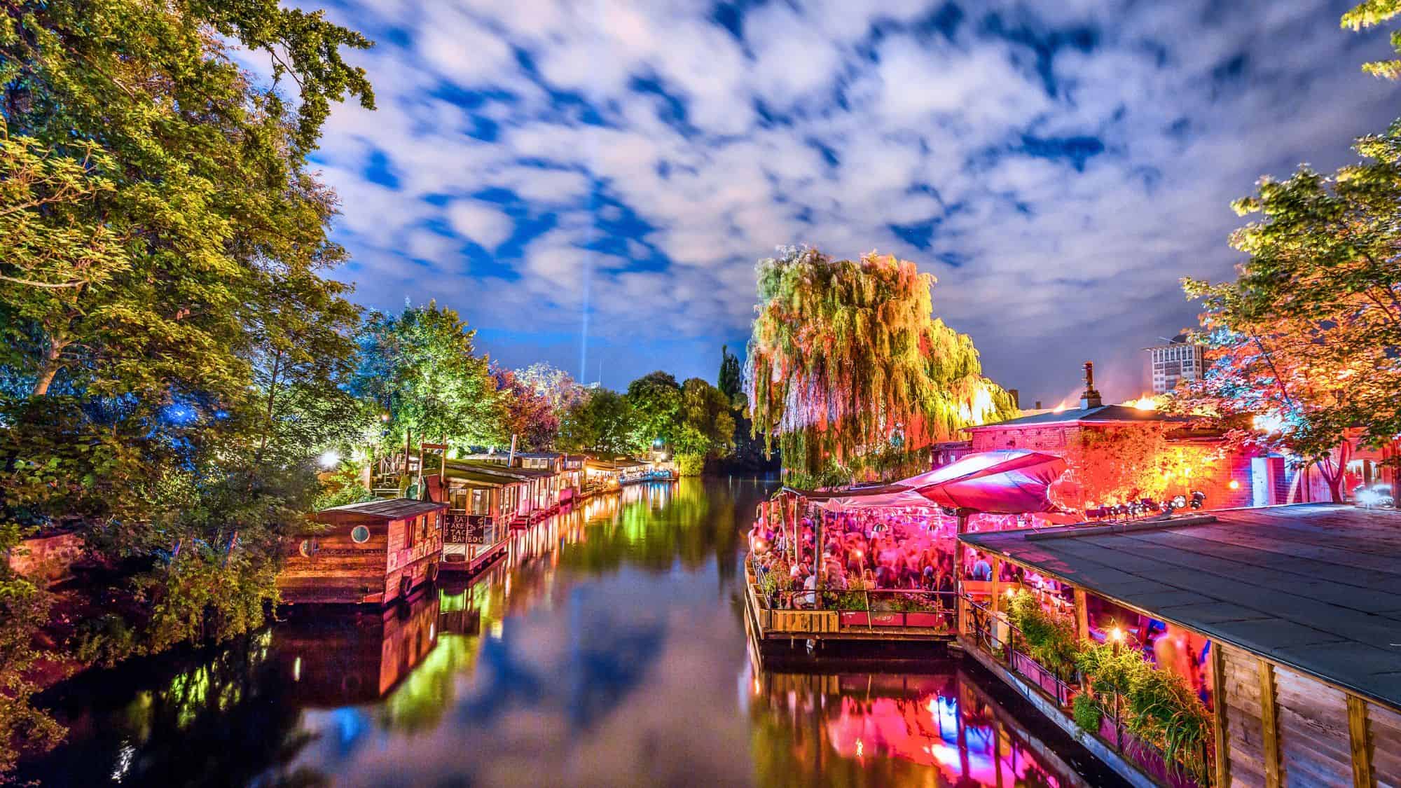 A lively crowd gathers at a brightly lit riverside bar in Berlin, Germany, where colorful lights reflect off the calm canal surrounded by trees and houseboats.