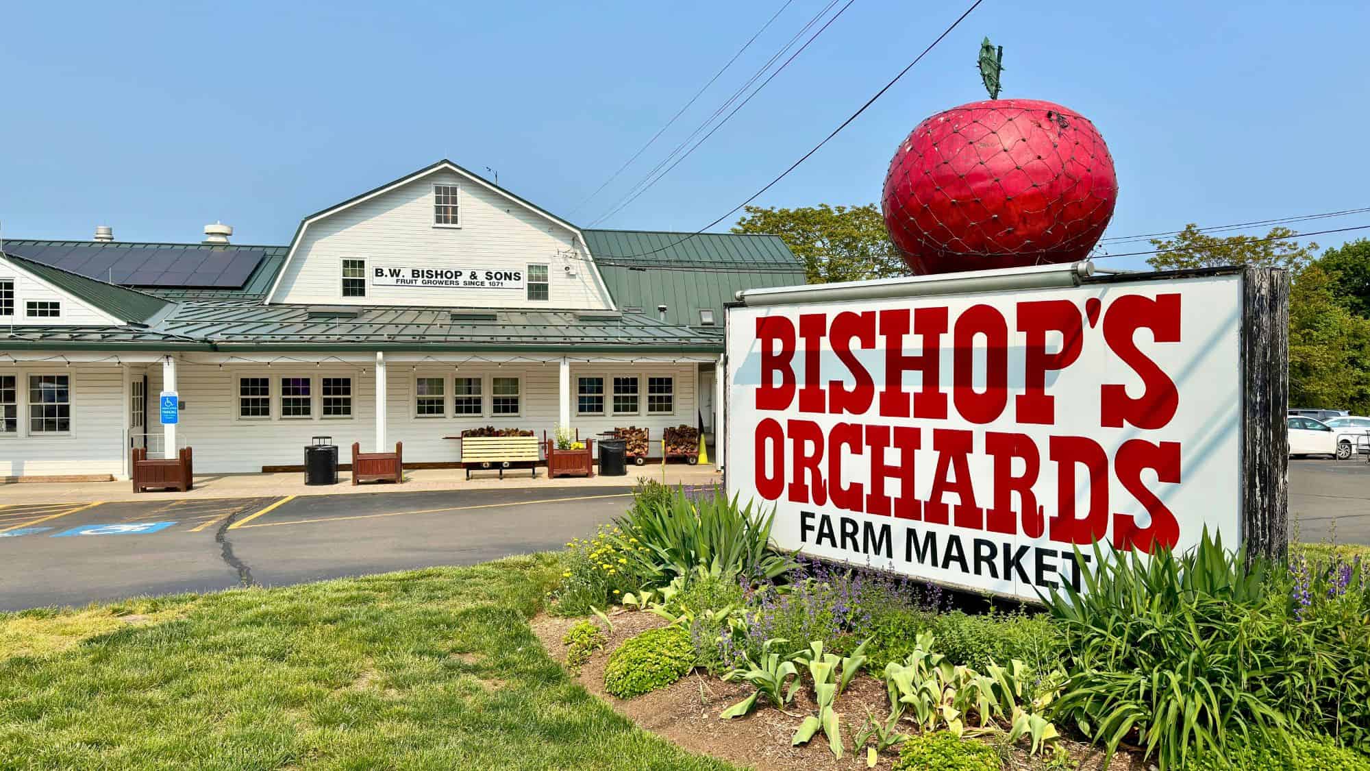 A large red sign with a giant apple sculpture reads “Bishop’s Orchards Farm Market” in front of a white building with green roofing and solar panels on a clear day.