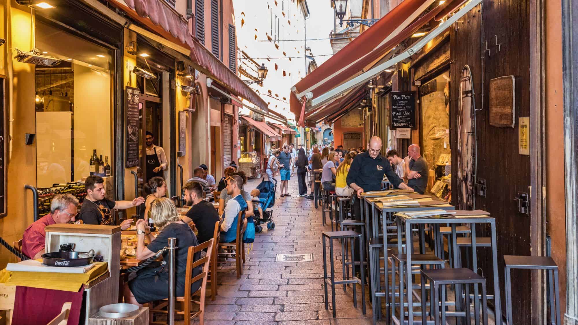 A bustling narrow alley in Italy is lined with outdoor restaurant tables, where people eat, drink, and chat under red awnings in warm afternoon light.