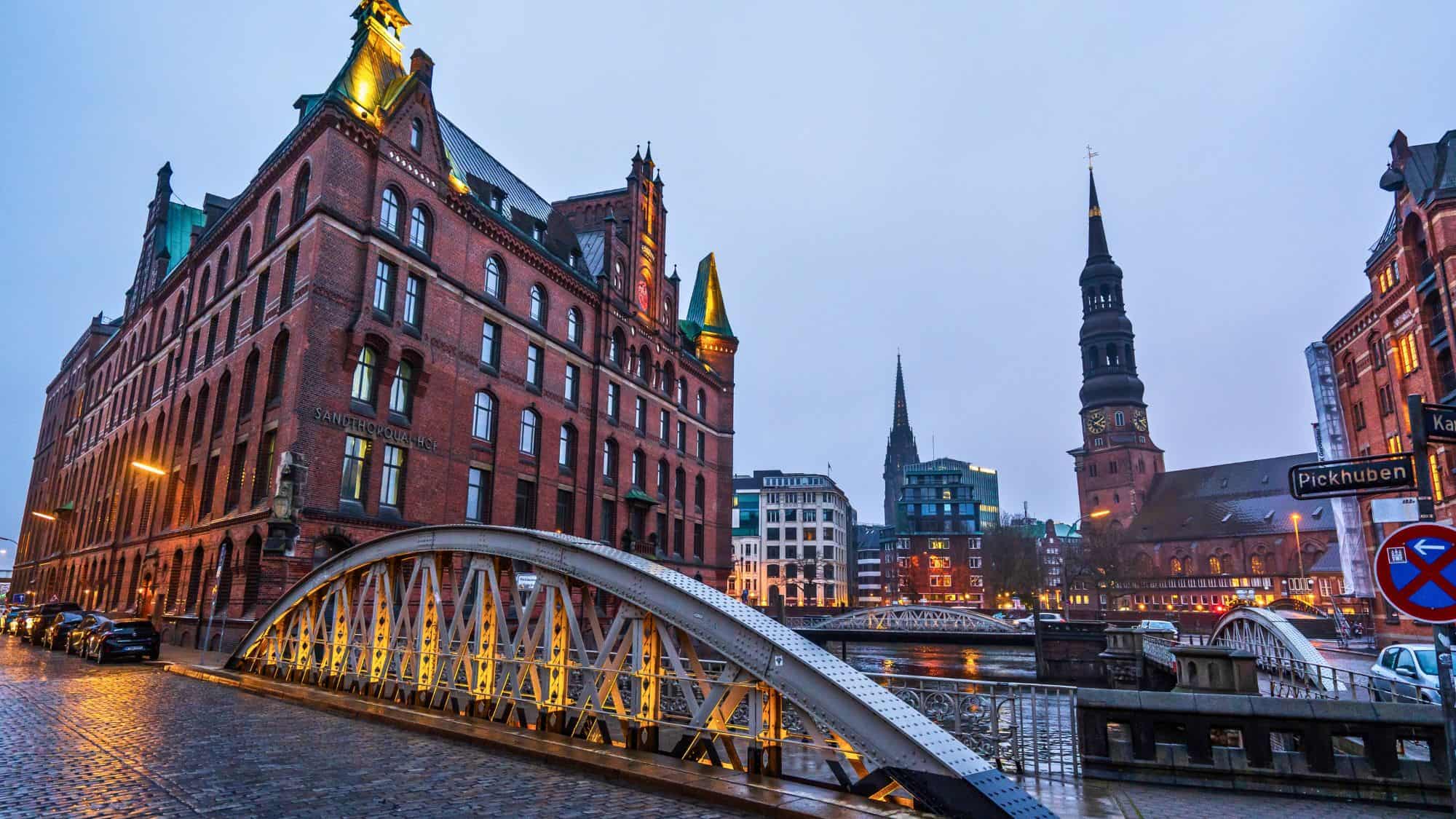 Historic red-brick warehouses and spired churches line the cobblestone streets and iron bridges of Hamburg’s Speicherstadt district during a misty evening.