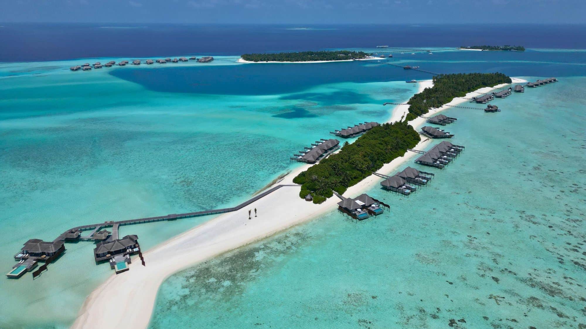 Aerial view of luxury overwater villas stretching along white sandbars and turquoise lagoons in the Maldives, surrounded by vivid coral reefs and deep blue ocean.