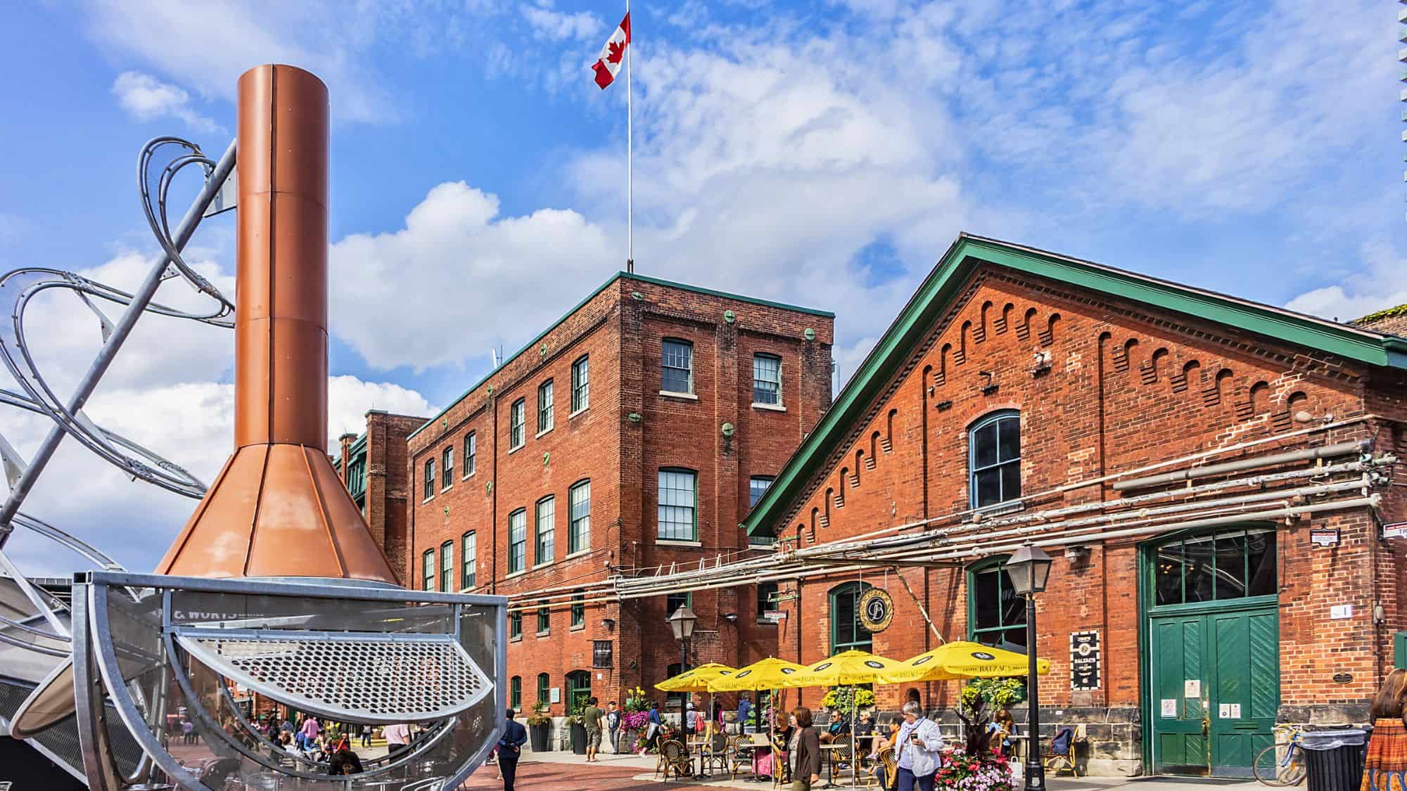 Red-brick industrial buildings with green-trimmed windows surround a sunny courtyard in Toronto’s Distillery District, featuring yellow patio umbrellas, visitors strolling, and a large copper brewing sculpture in the foreground under a Canadian flag.