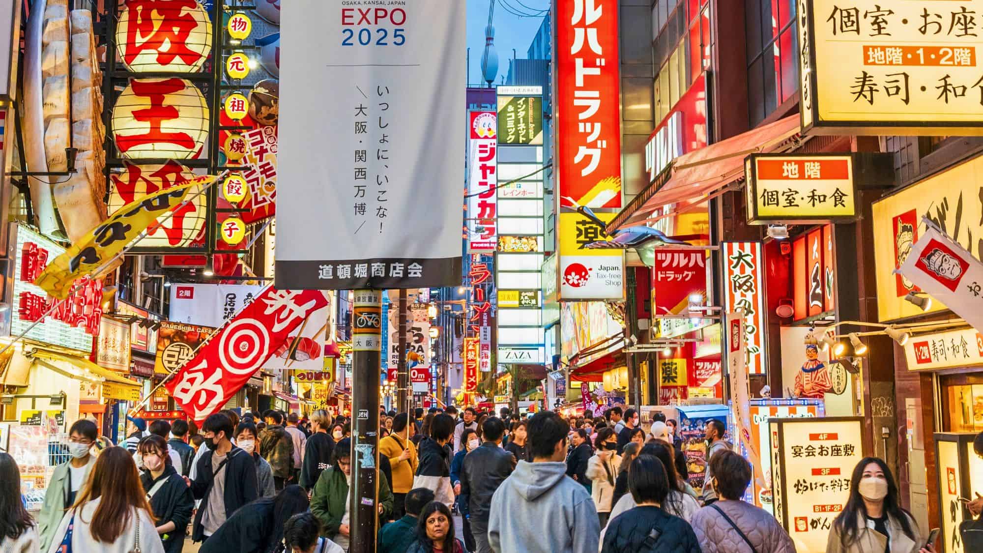 A lively street in Osaka’s Dotonbori district packed with people and lit by a dense array of colorful signs, lanterns, and advertisements in Japanese.