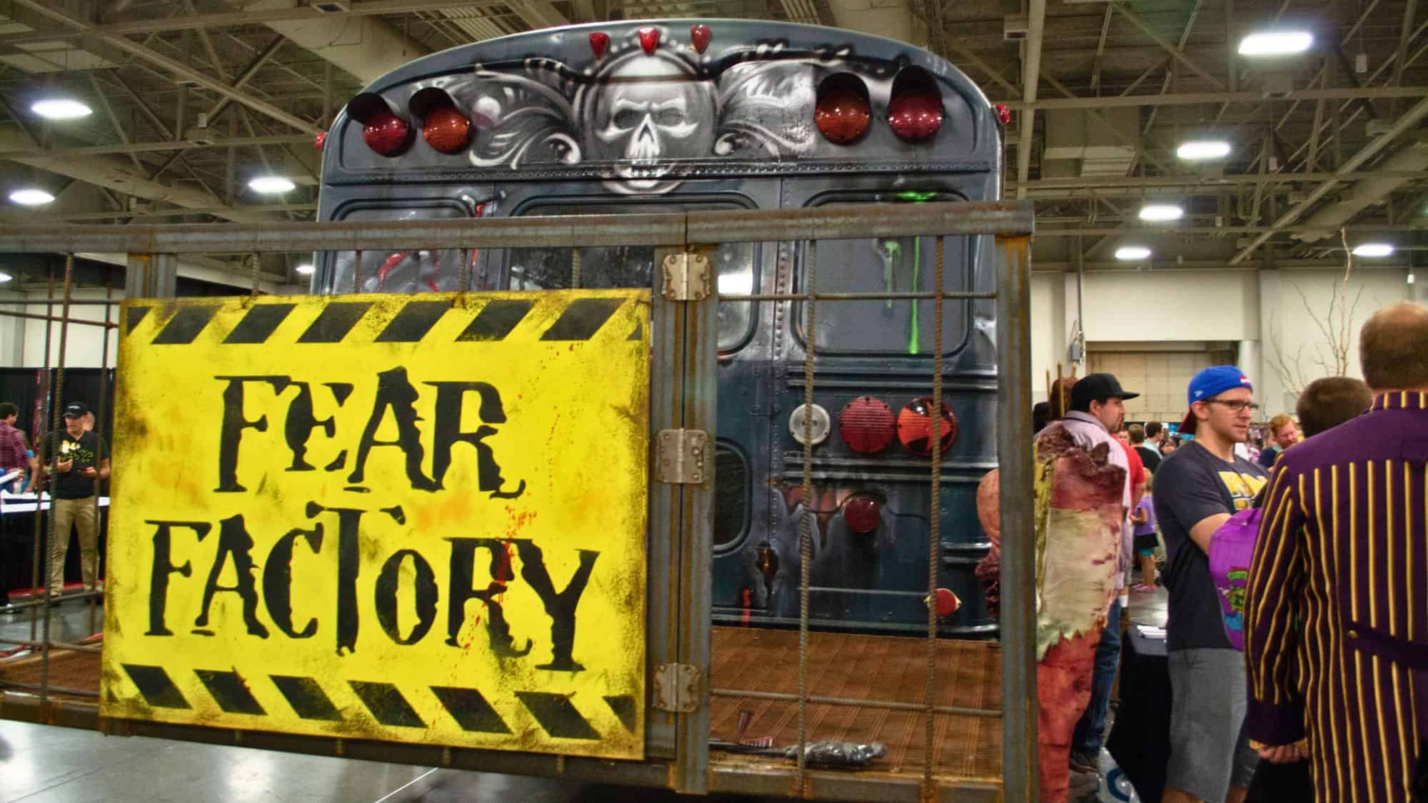 A spooky convention display features a repurposed bus with skull-themed graffiti and a bright yellow "Fear Factory" sign in front, surrounded by costumed attendees.
