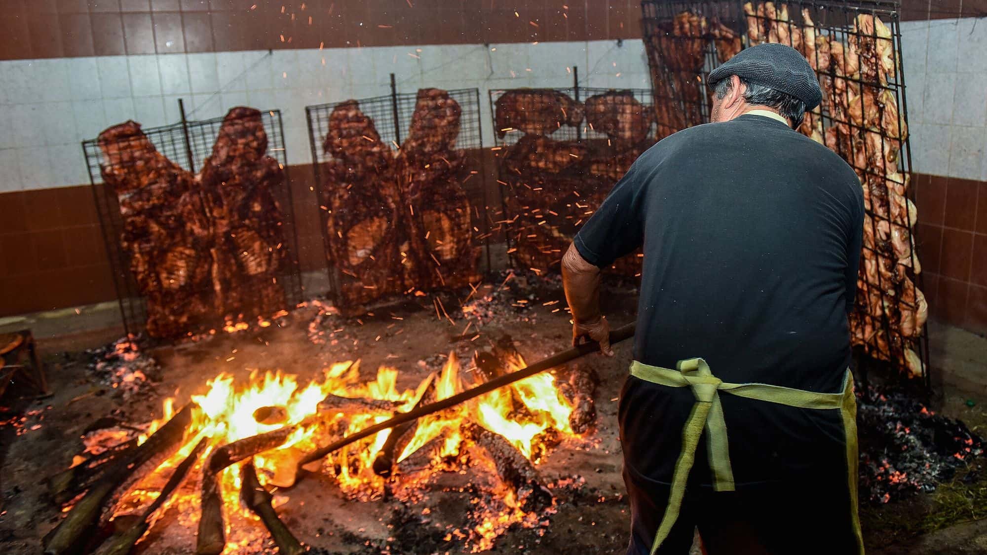 A man in a black shirt and cap tends to a traditional Argentine asado, where slabs of meat are slow-roasted upright around an open wood fire.