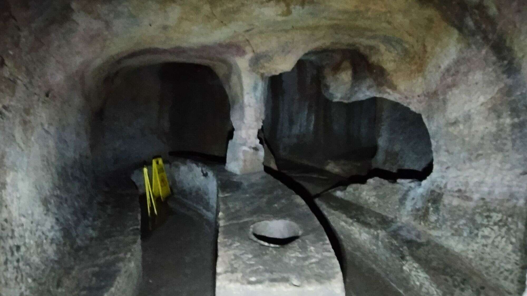 A dim interior of the Glimerton Cove shows a narrow, hand-dug corridor with smooth rock walls and a stone platform marked by a caution sign.