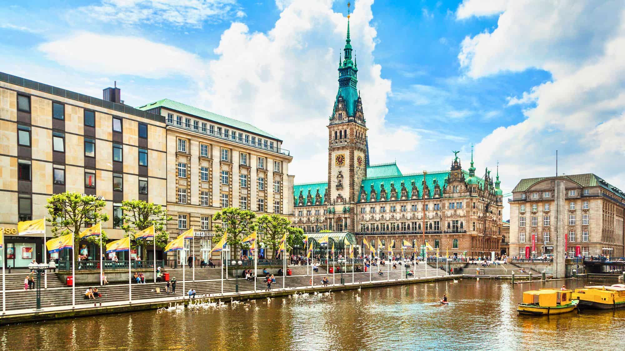 The ornate Hamburg Rathaus with its green copper roof towers over a canal bustling with boats and lined by lively public steps and modern buildings.