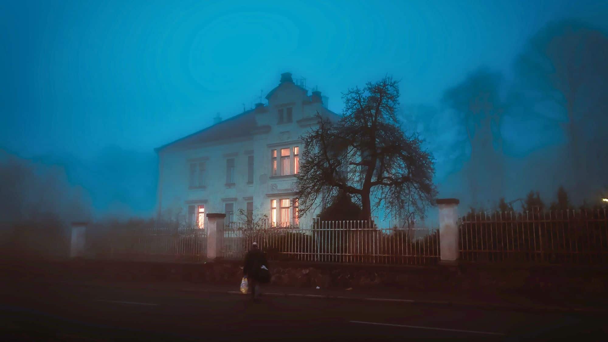 A solitary figure walks past a gated, two-story house glowing warmly from within, shrouded in thick blue fog and framed by a bare, eerie tree.