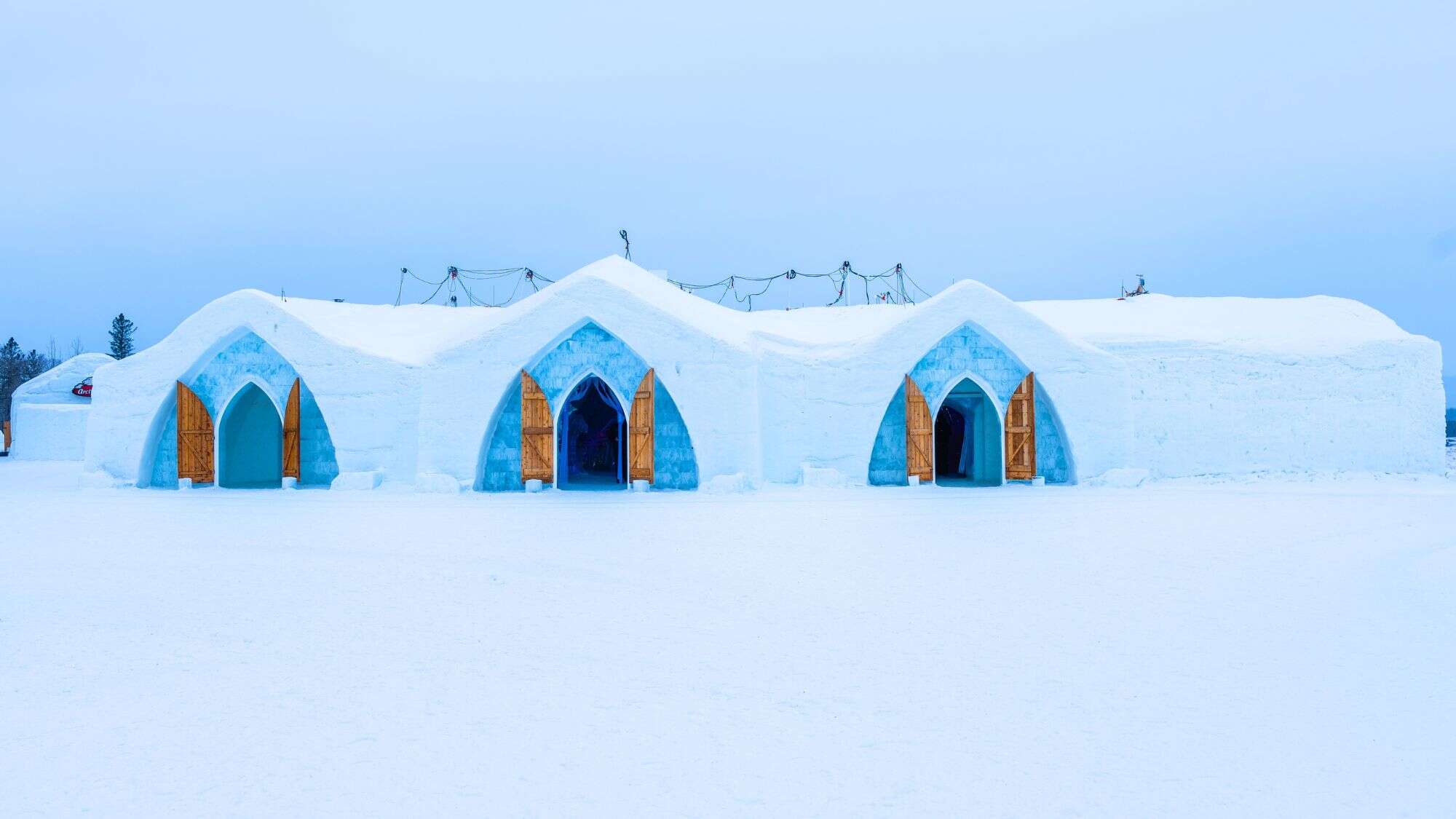 A symmetrical structure made entirely of snow and ice stands against a pale sky, with pointed arch doorways and warm wooden doors at the entrance to a winter hotel.