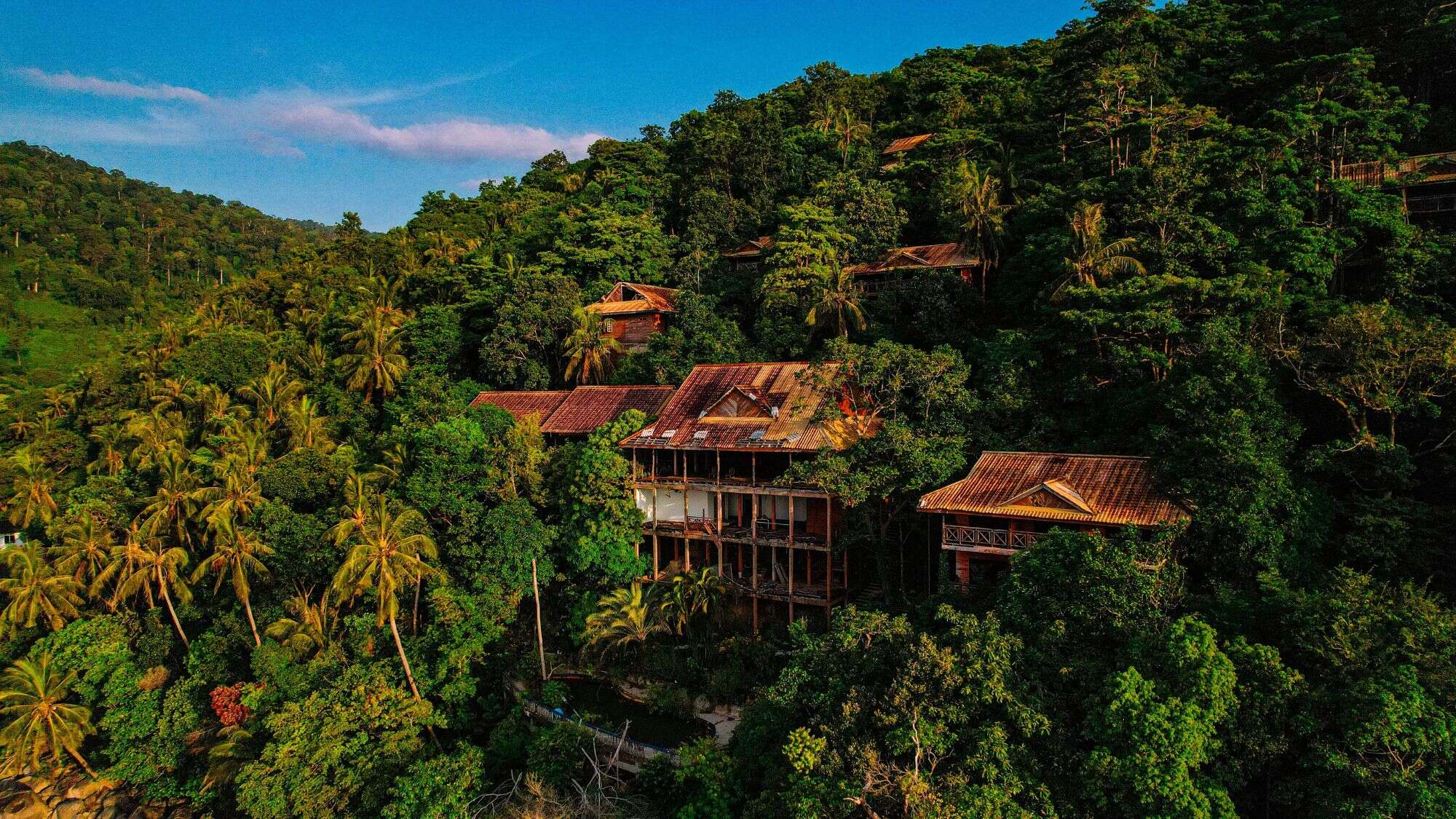 Wooden villas with rust-colored roofs are nestled deep in lush, green jungle hills, blending into the dense tropical forest canopy.