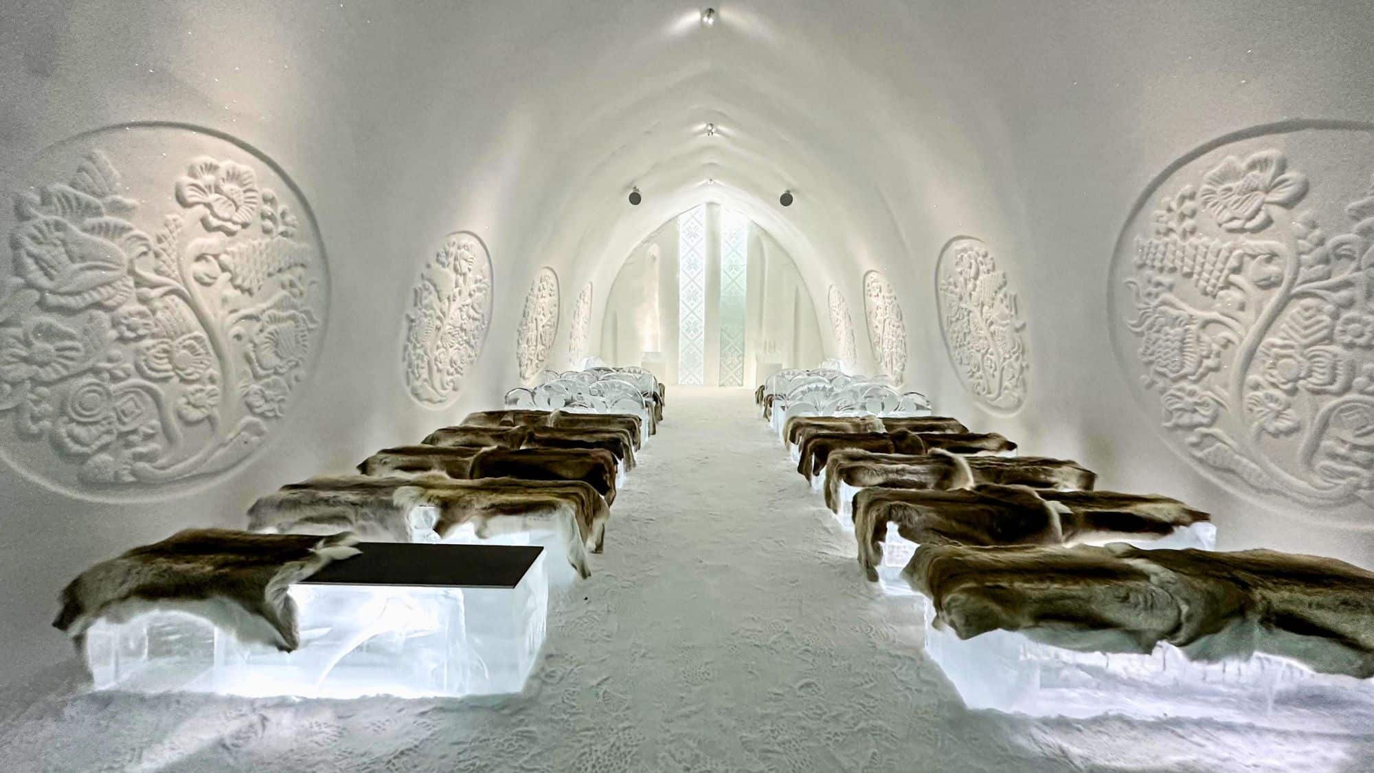 A softly lit chapel carved from snow and ice features intricate floral reliefs on the walls, with benches topped in furs and a glowing altar at the far end.