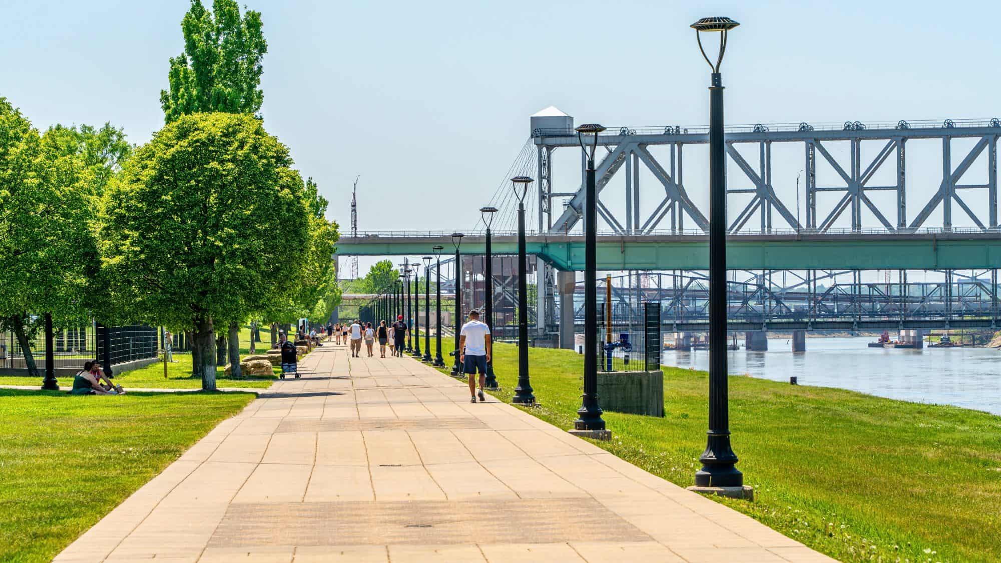 A sunny riverside path lined with trees and lampposts runs beside the St. Lawrence River in Montreal, with large steel bridges spanning the water in the distance.
