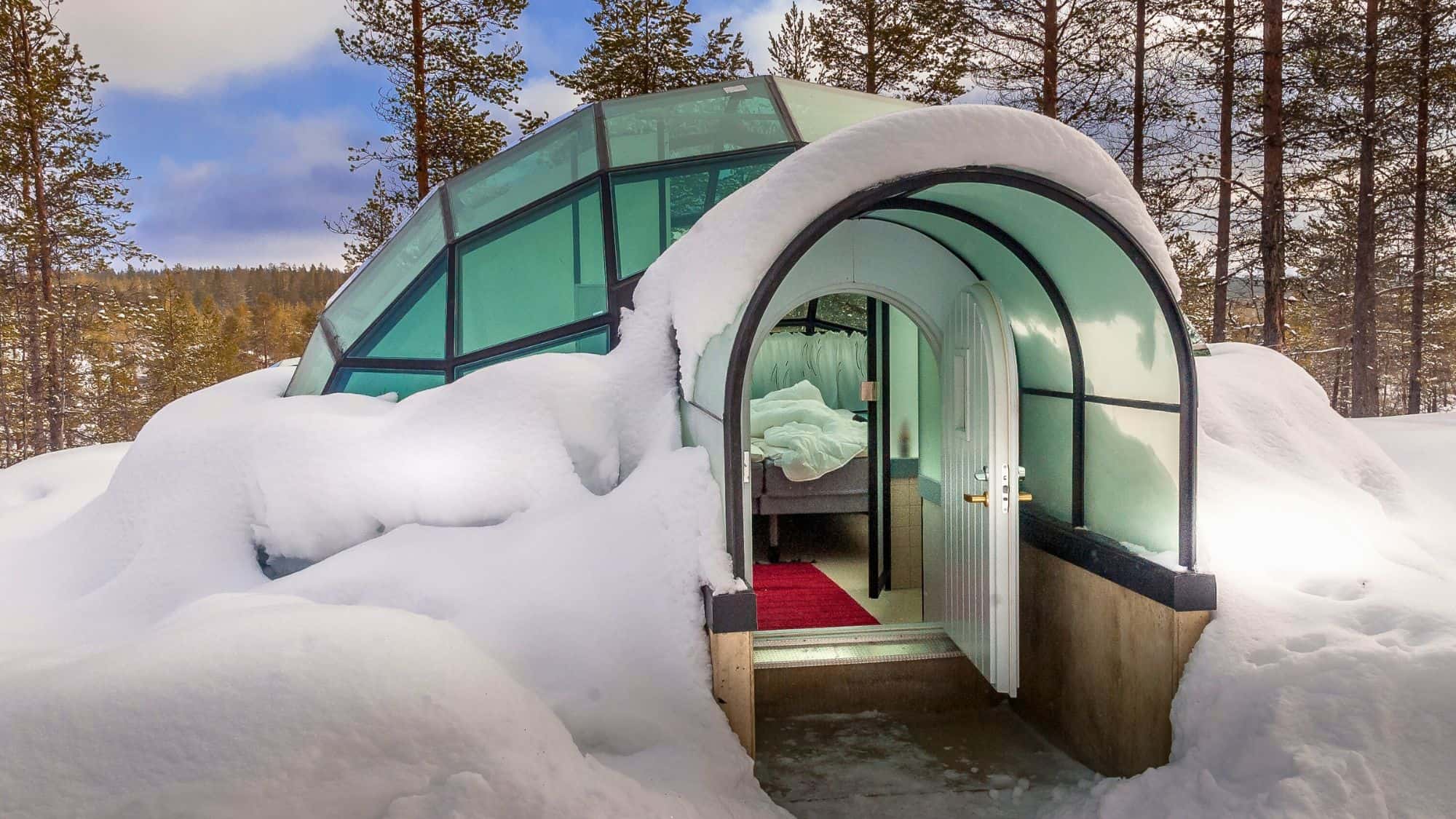 A glass-domed igloo stands surrounded by heavy snow in a Finnish forest, offering a cozy, transparent shelter for viewing the winter landscape and northern lights.