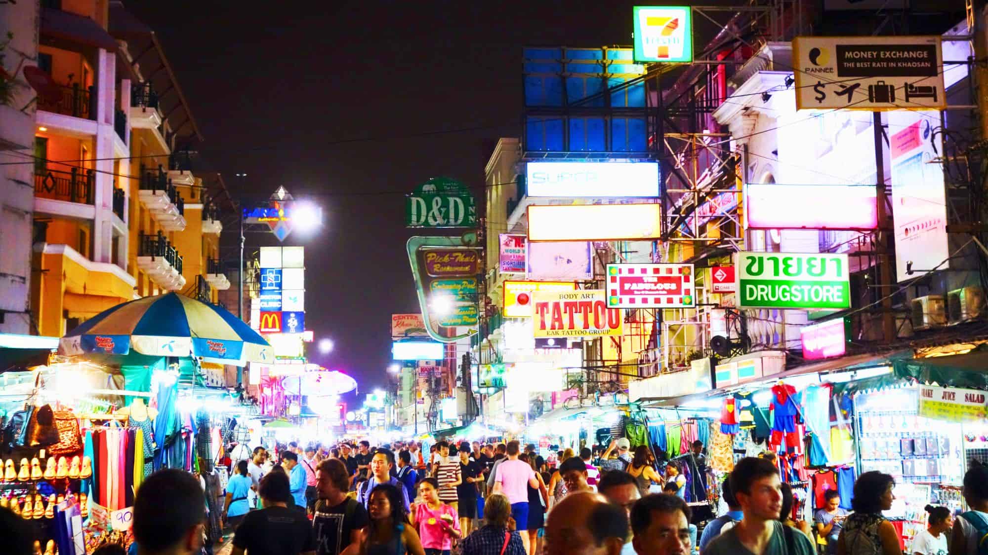 Bangkok’s Khao San Road is packed with tourists and locals beneath a jungle of neon signs, food stalls, and street vendors, creating a chaotic and energetic nighttime atmosphere.