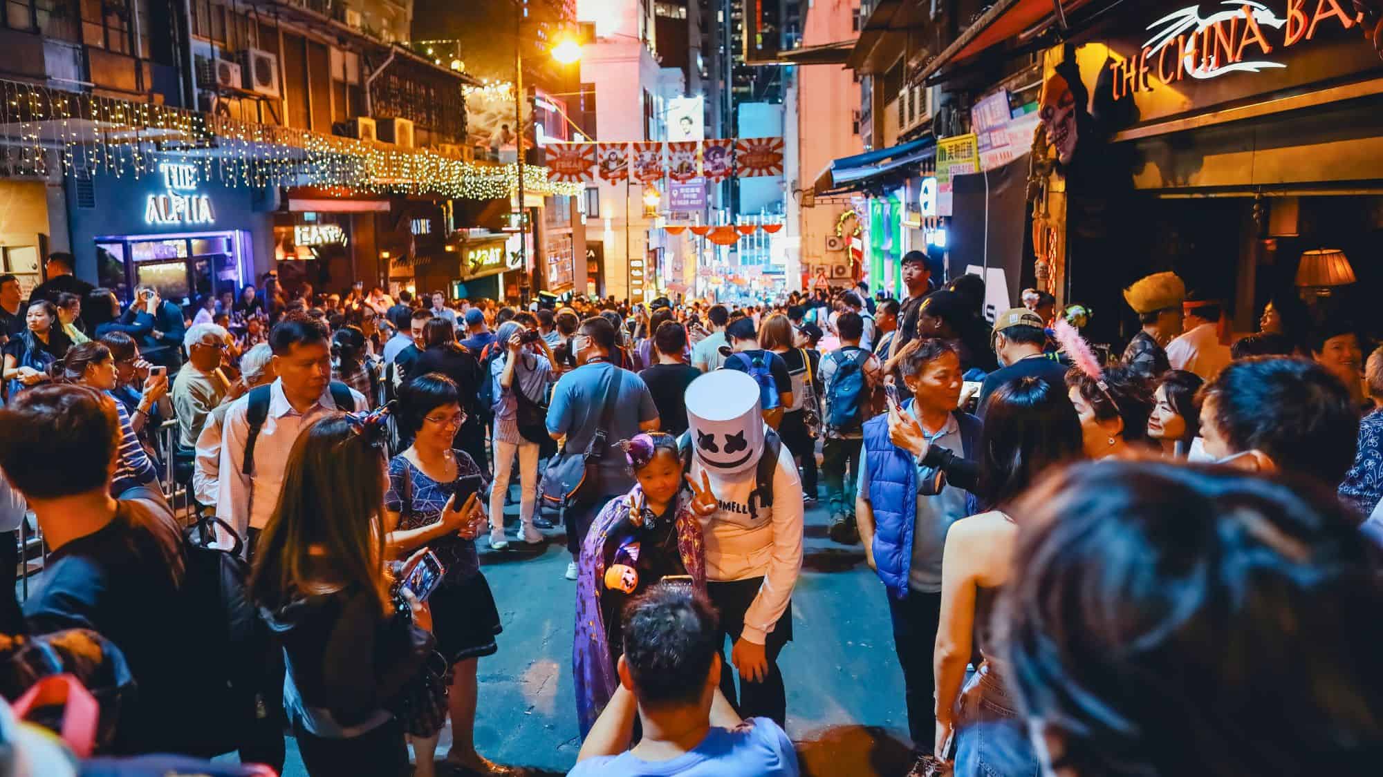 A dense crowd fills the narrow, bar-lined streets of Lan Kwai Fong in Hong Kong, many dressed in costumes for Halloween under festive lights and neon signs.