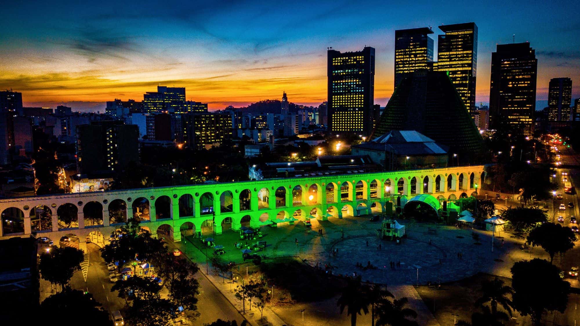 The Arcos da Lapa in Rio de Janeiro are illuminated green against a vivid sunset sky, surrounded by high-rises and the conical silhouette of the Metropolitan Cathedral.