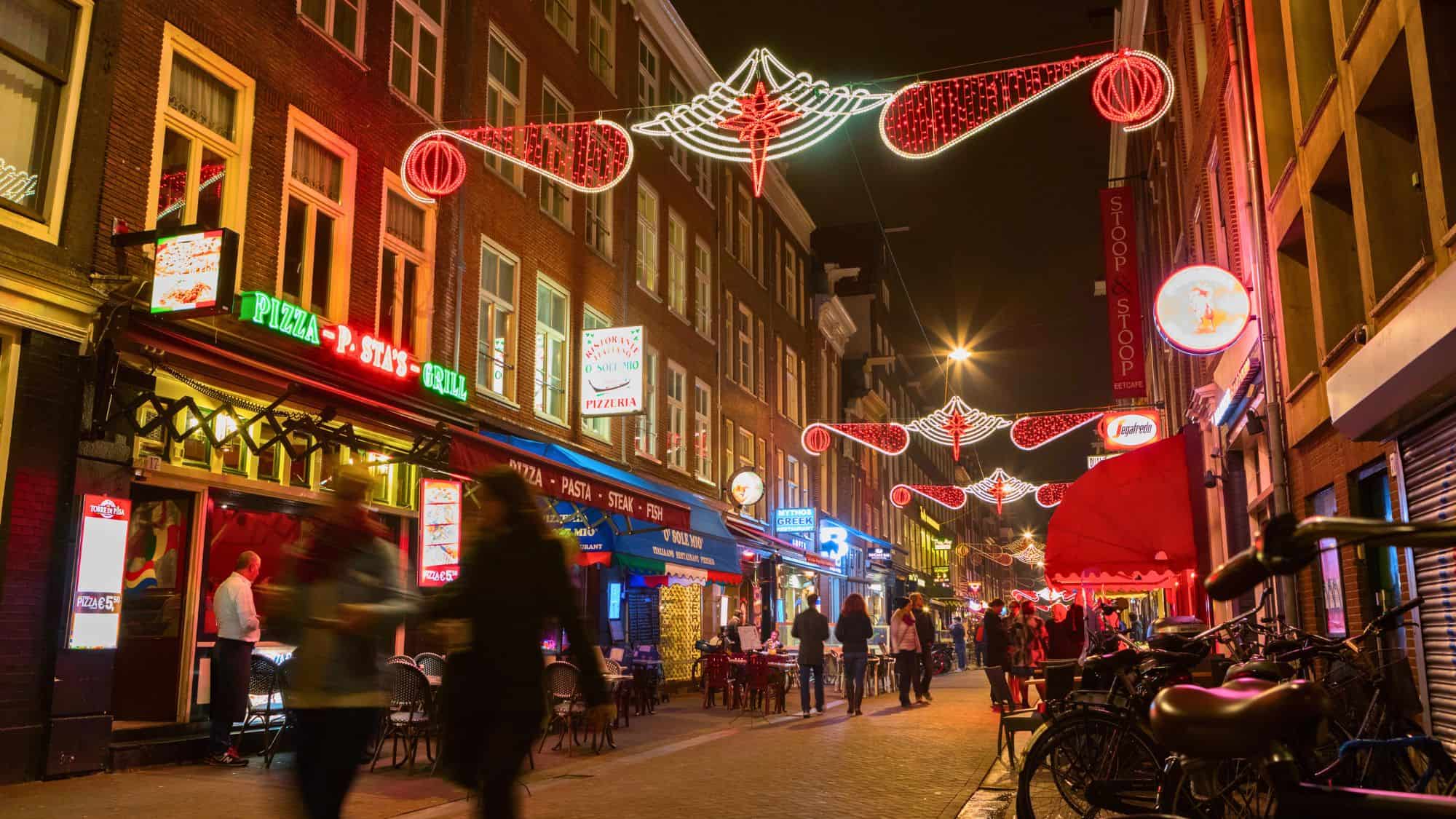 A warmly lit Amsterdam street glows under holiday lights with people walking past pizzerias and cafés, showcasing a cozy nightlife scene with red-brick charm.