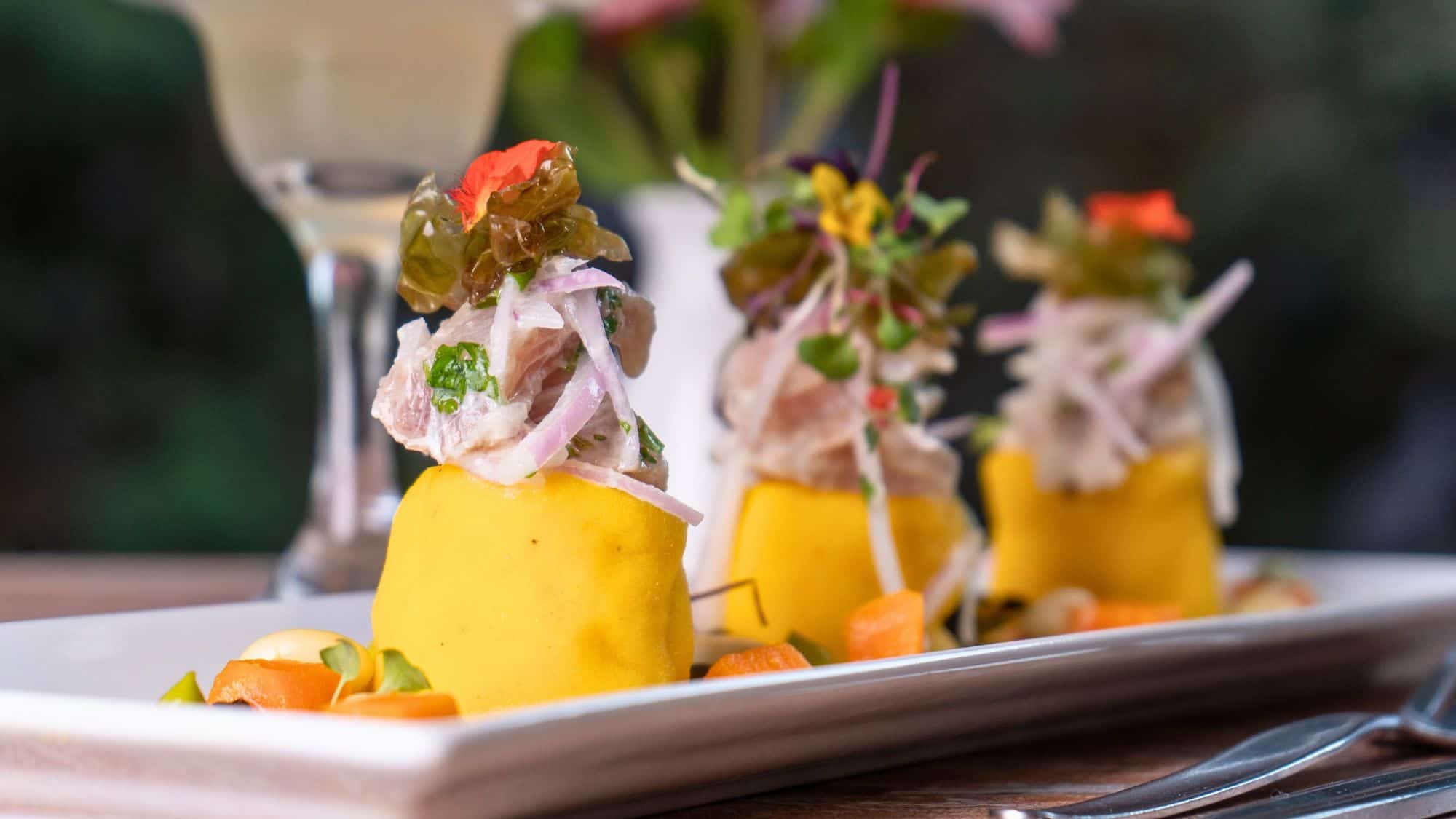 A gourmet plate of causa rellena features vibrant yellow potato stacks topped with seafood, onions, and edible flowers on a white dish.