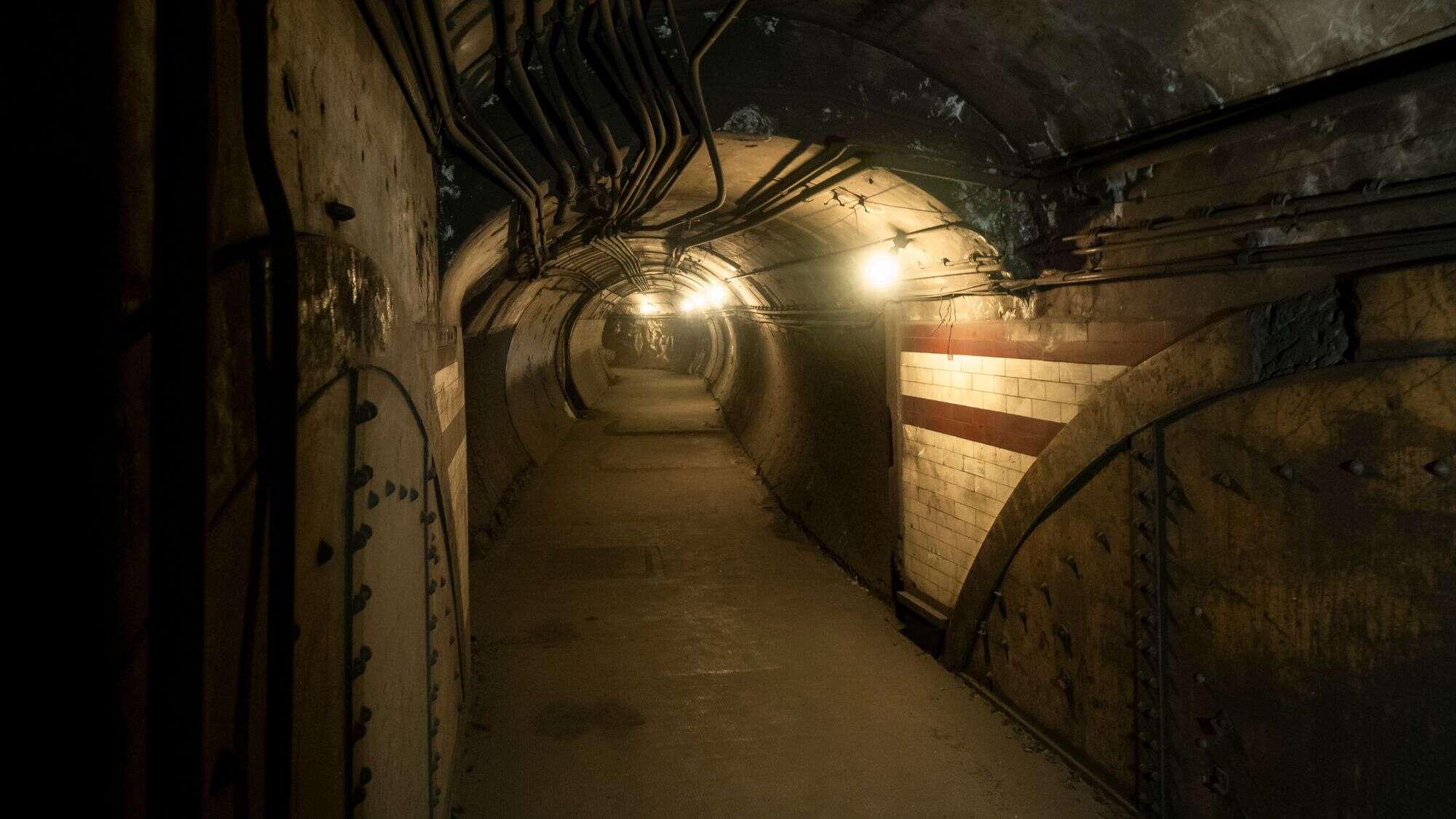 A dim, grimy underground tunnel with exposed pipes and aging tiled walls curves into the distance, lit by exposed yellow bulbs hanging from the ceiling.