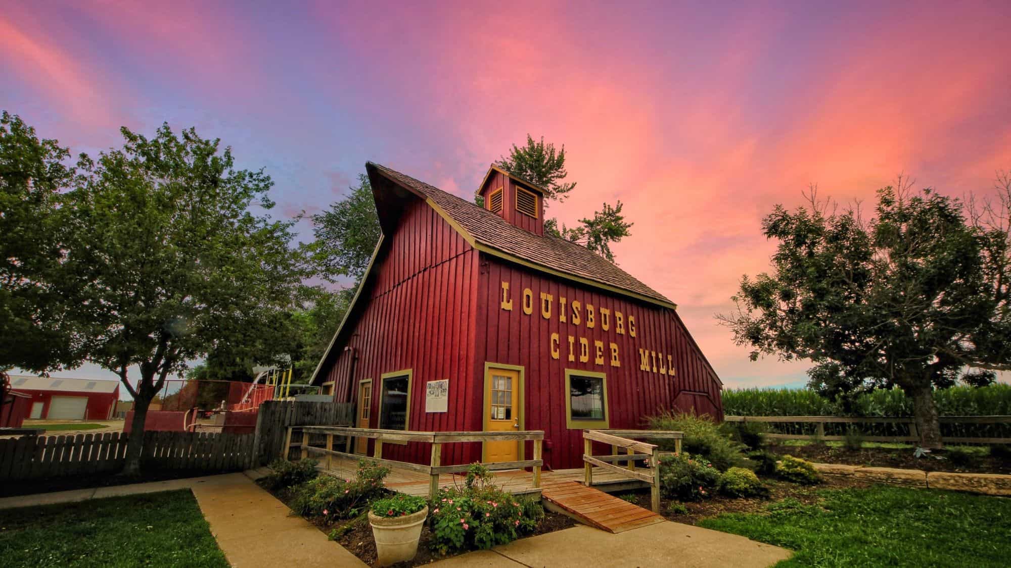 A rustic red barn labeled “Louisburg Cider Mill” stands beneath a vibrant pink and orange sunset sky, surrounded by trees, a wooden ramp, and garden flowers.