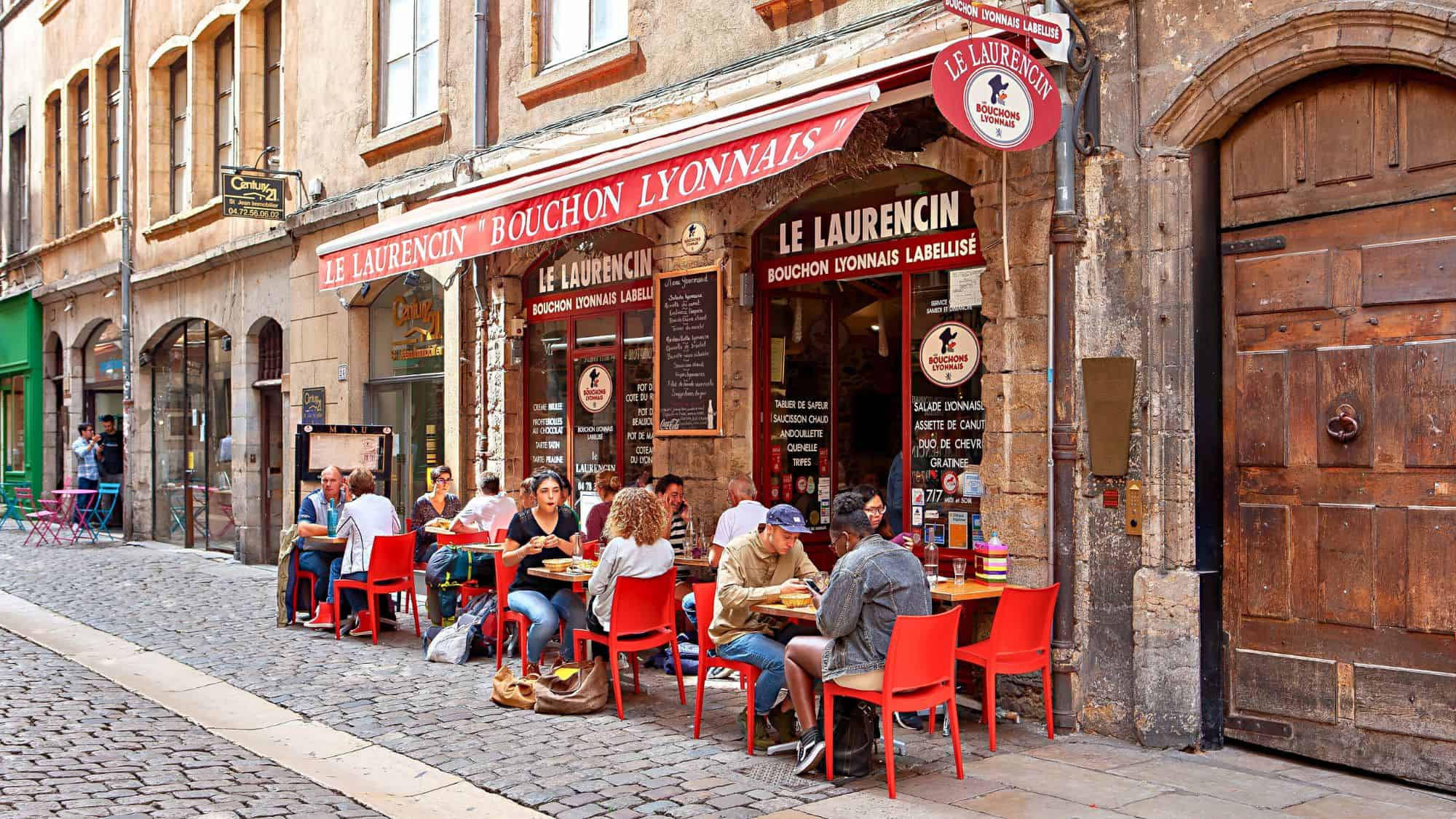 Diners enjoy traditional Lyonnaise cuisine at red tables outside "Le Laurencin Bouchon Lyonnais" on a cobblestone street in Lyon, France.