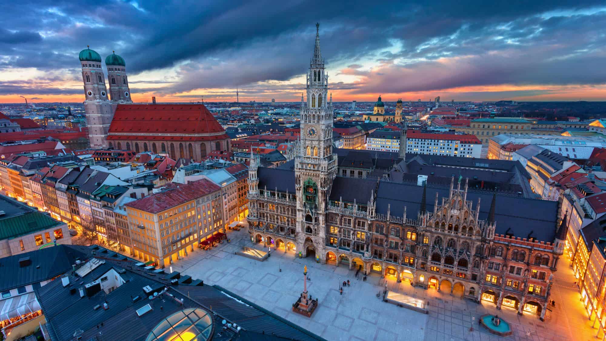 A sweeping view of Munich’s city center at sunset shows the Neues Rathaus and Frauenkirche rising over the rooftops, with soft lighting spilling into the square.
