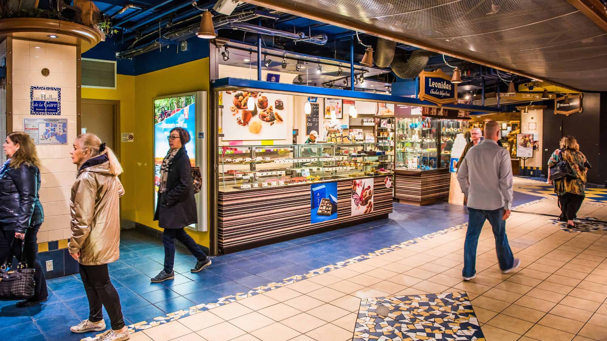 A vibrant underground shopping area in Montreal features a Belgian chocolate shop and glass display cases, with shoppers browsing under mosaic-tiled floors.