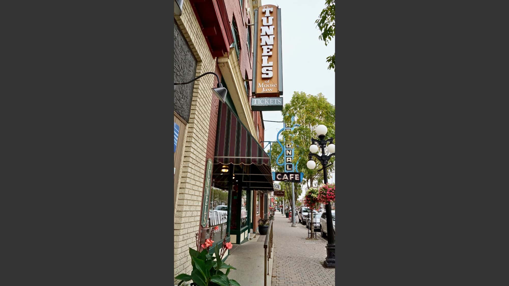 A sidewalk view of the Tunnels of Moose Jaw entrance in Saskatchewan, Canada, with vintage-style signage and storefronts along a historic brick street.
