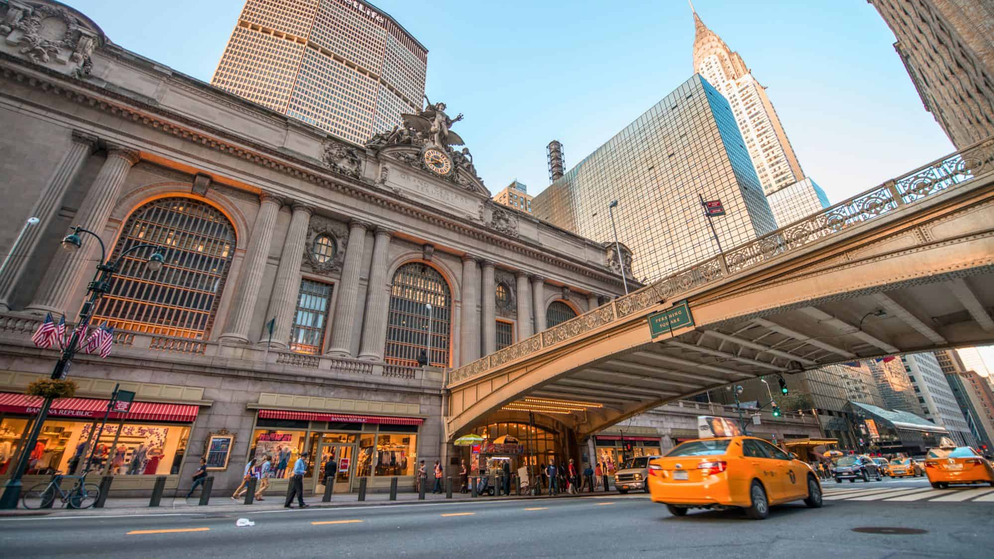 The exterior of Grand Central Terminal with its grand arched windows and yellow taxis passing in front, set against tall skyscrapers.