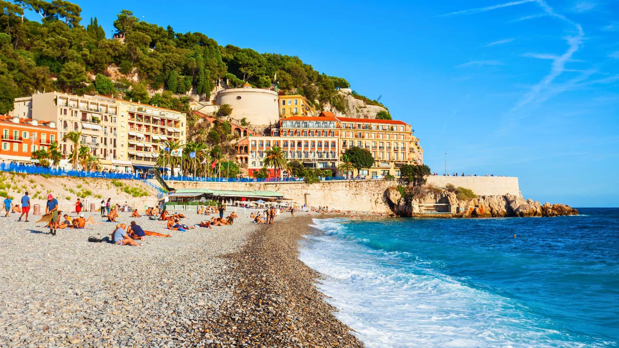 Sunbathers relax on the pebbled beach of Nice with pastel buildings and lush greenery rising above the shoreline.