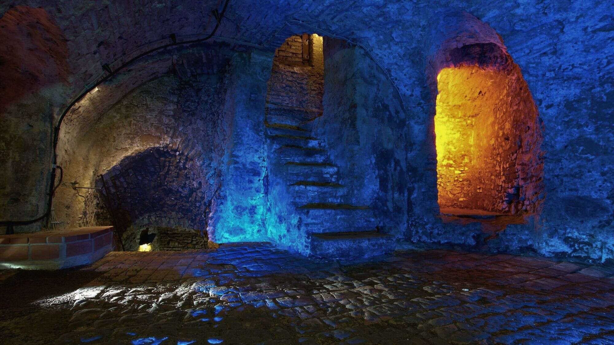 Stone staircases and arched rooms of the Edinburgh Vaults are illuminated in dramatic blue and orange lighting, casting colorful reflections on the damp stone floor.