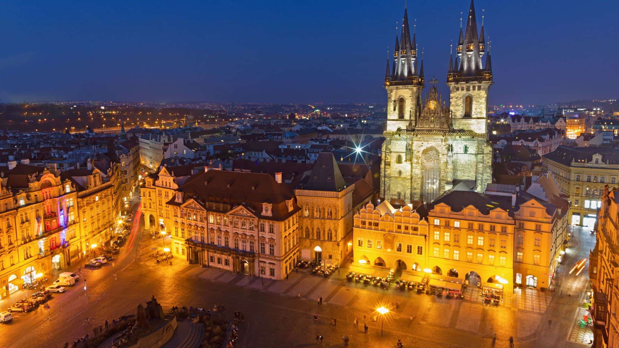 The Gothic spires of the Church of Our Lady before Týn rise above Prague’s Old Town Square, which is bathed in golden light with surrounding Baroque buildings.