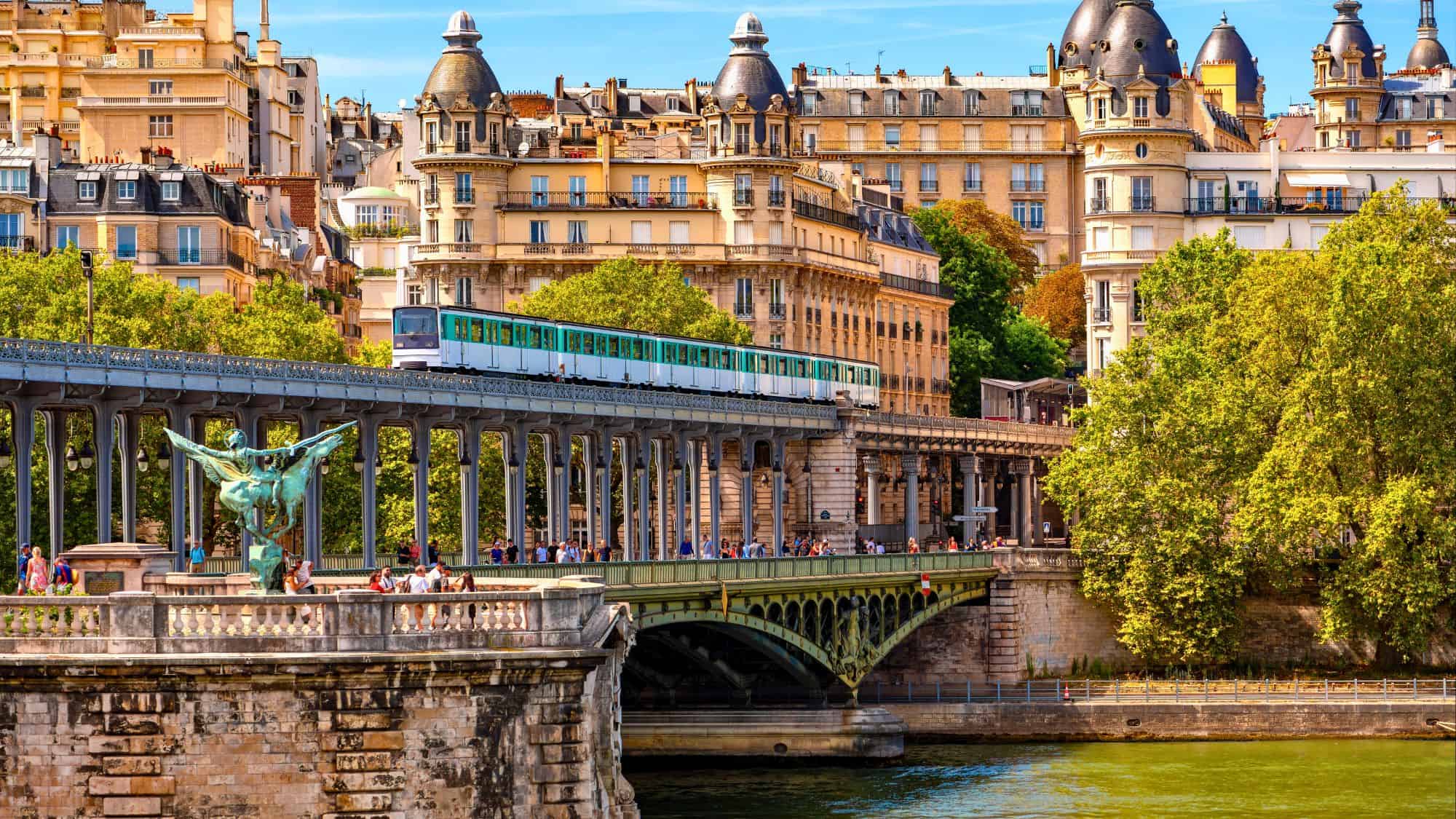 A Paris Métro train crosses a historic iron bridge over the Seine, with ornate statues and Haussmann-style buildings in the background.
