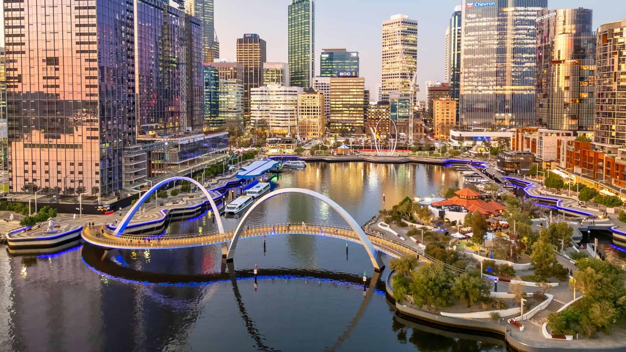 Curved pedestrian bridges and modern boardwalks wrap around the calm inlet of Elizabeth Quay in Perth, Australia, with high-rise towers glowing at sunset.