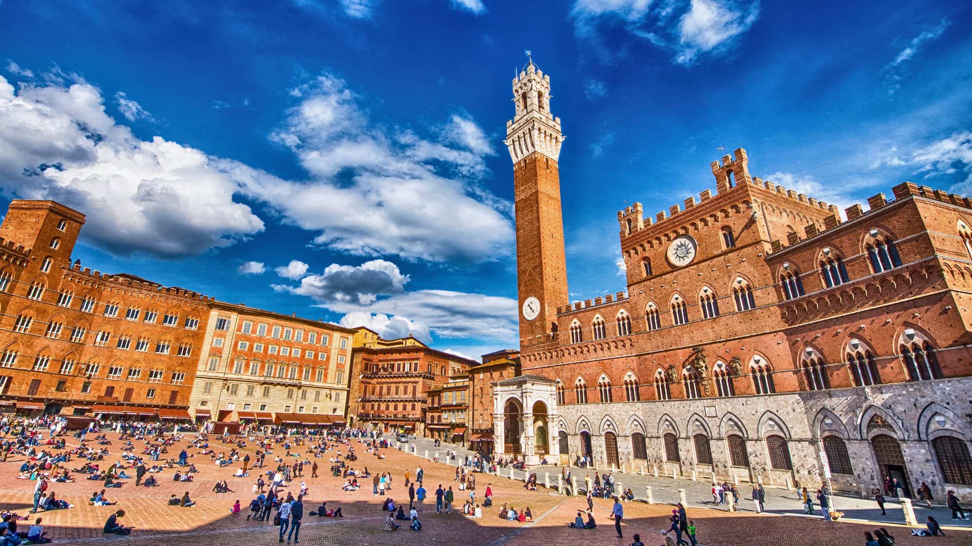 The shell-shaped square in Siena is filled with people lounging on brick pavement, framed by medieval buildings and the tall Torre del Mangia under a vivid blue sky.