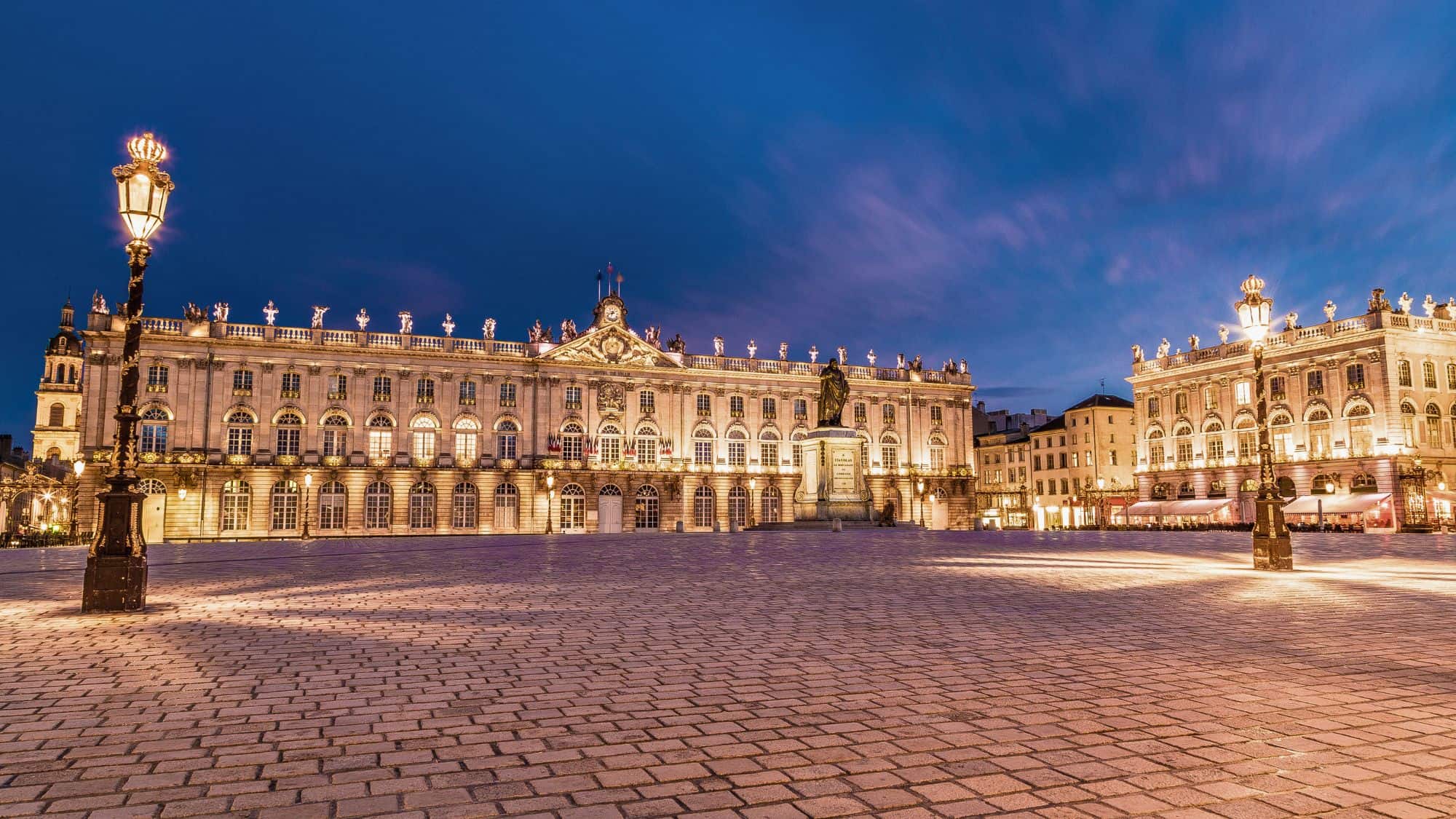 This elegant French square is illuminated with golden light, showcasing the Baroque architecture, ornate ironwork, and central statue of King Stanislas under a twilight sky.