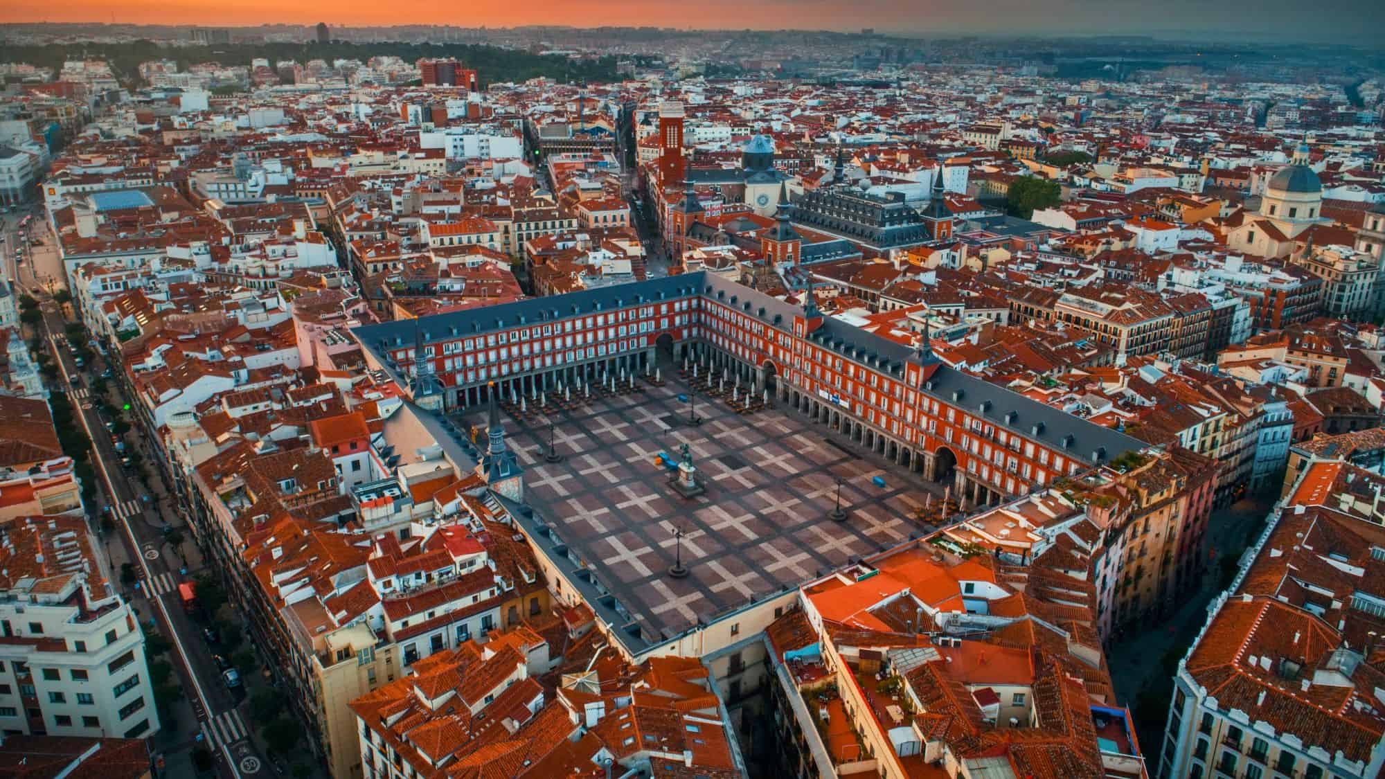 An aerial view of Madrid shows the symmetrical Plaza Mayor enclosed by red-roofed buildings, glowing with the last light of sunset.