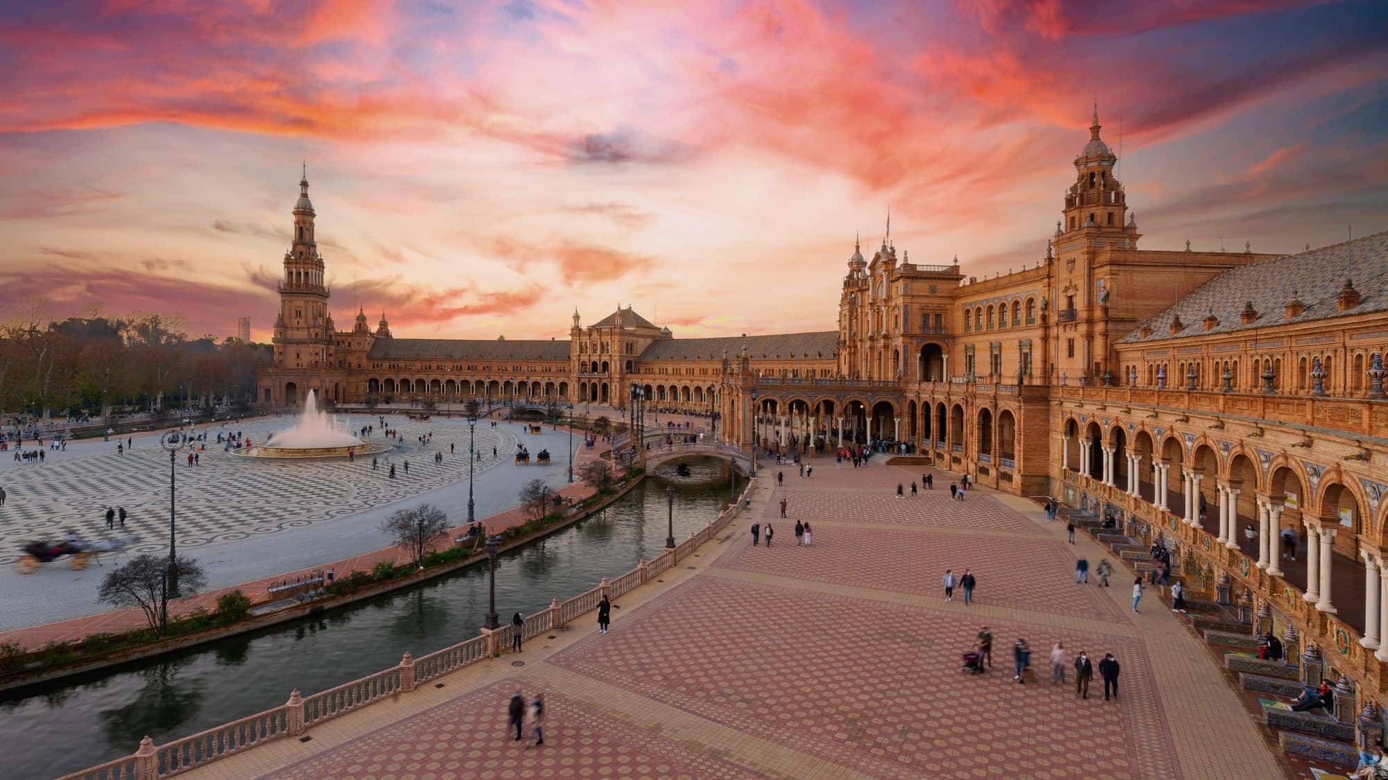 The grand semicircular plaza in Seville glows under a dramatic pink and orange sky, with crowds walking near the central fountain and ornate canal bridges.