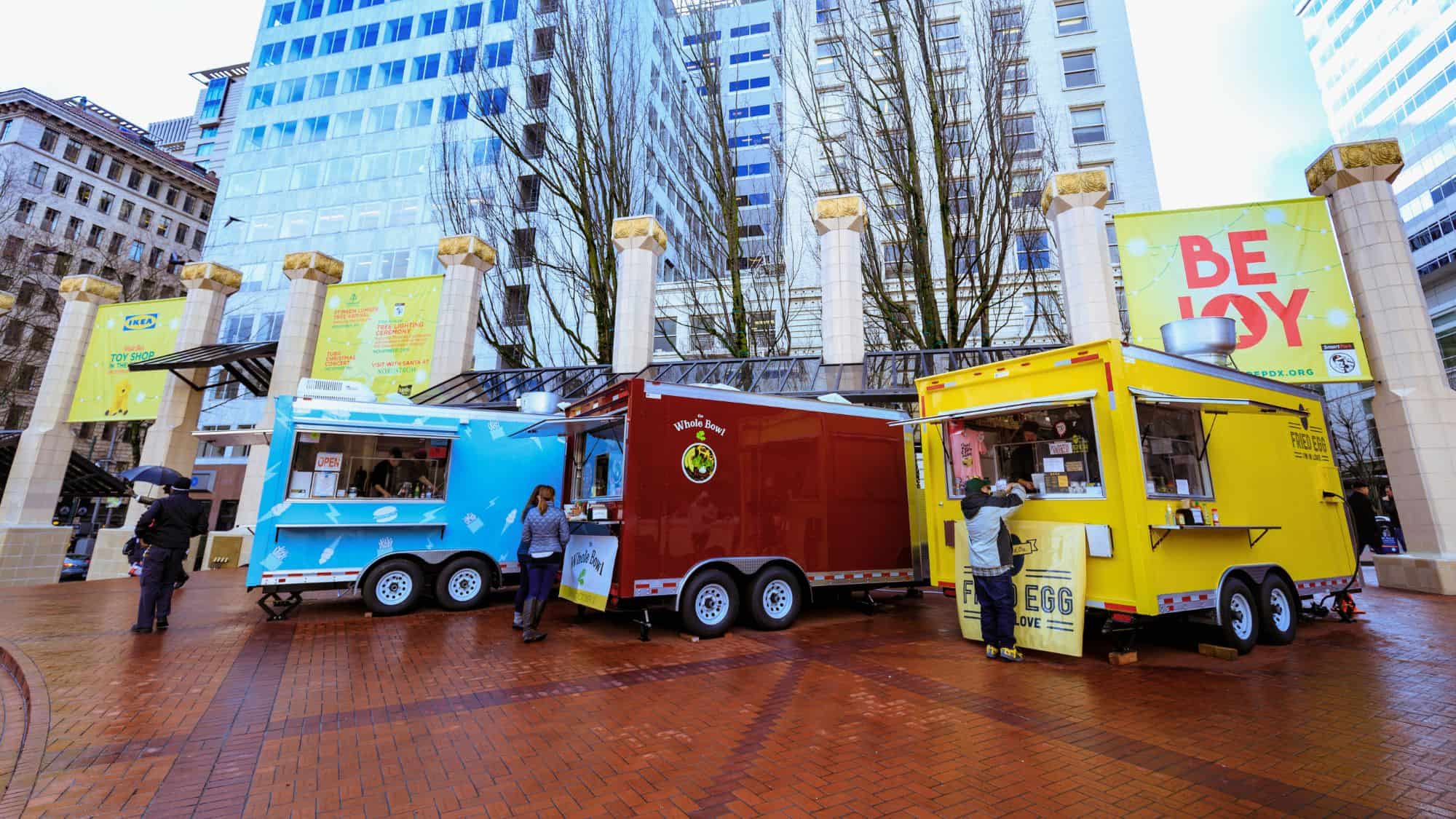 Three colorful food trucks—blue, red, and yellow—serve customers in a brick plaza in downtown Portland, Oregon, beneath tall buildings and winter trees.