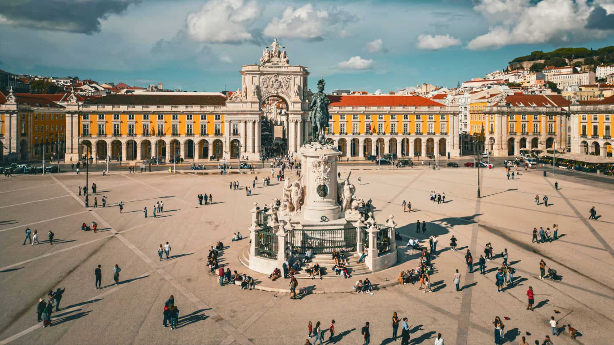 This vast square in Lisbon features the Arco da Rua Augusta and a statue of King José I, surrounded by yellow neoclassical buildings and bustling with people.