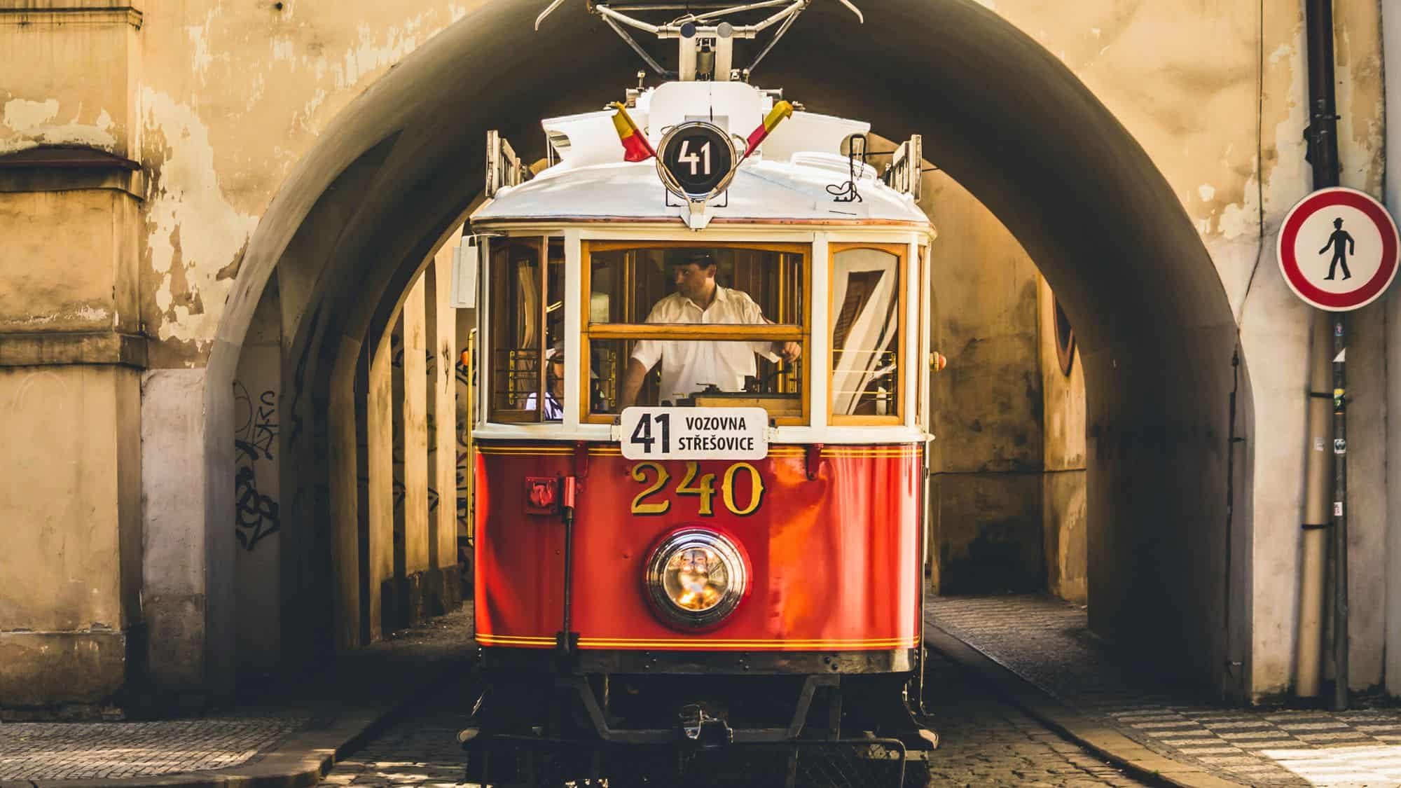 A historic red tram with gold accents passes under an old stone archway, with its driver visible inside.