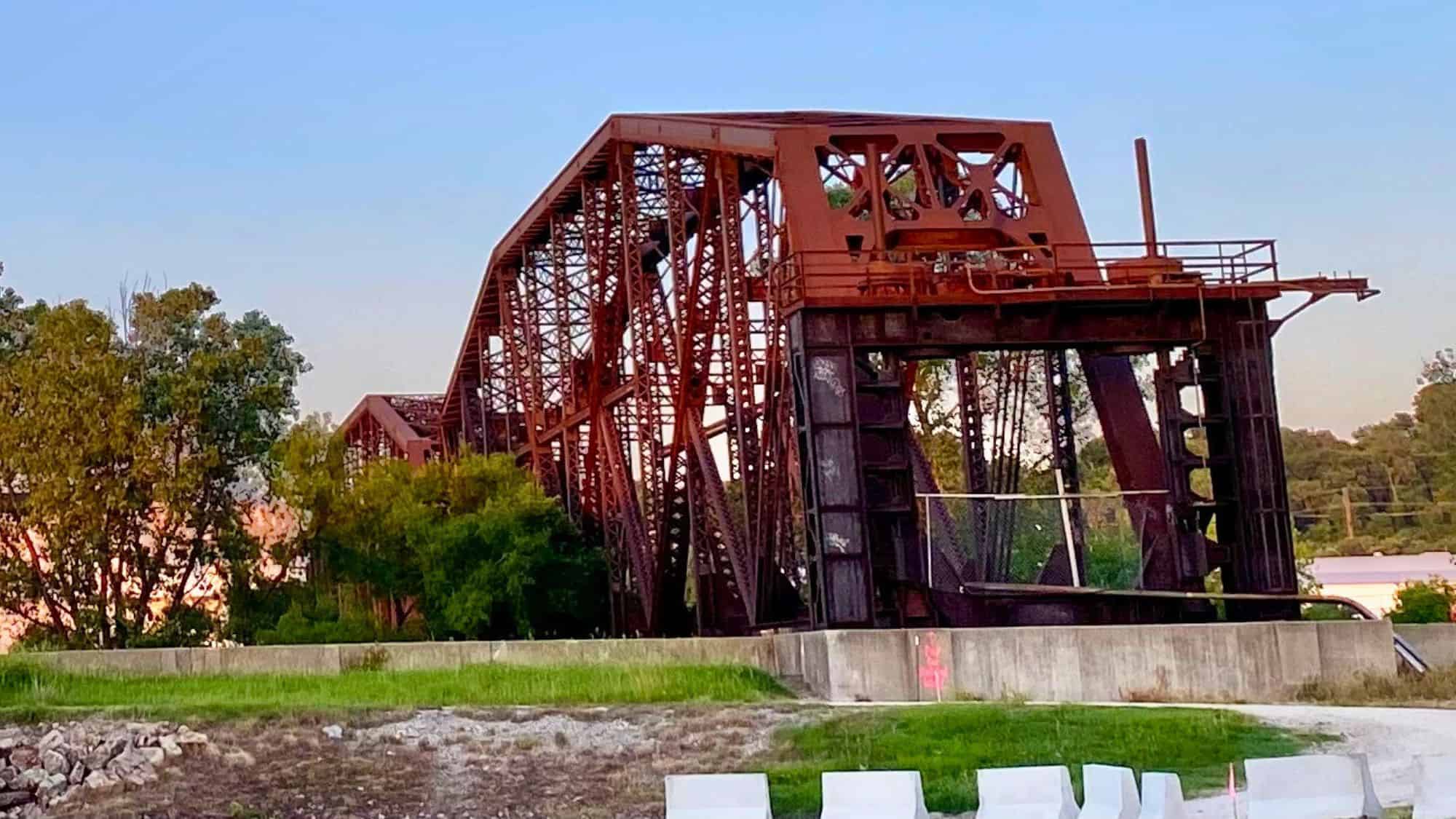 A weathered, rust-red railroad swing bridge stands partially open beside green trees and concrete embankments, showing signs of age and disuse.