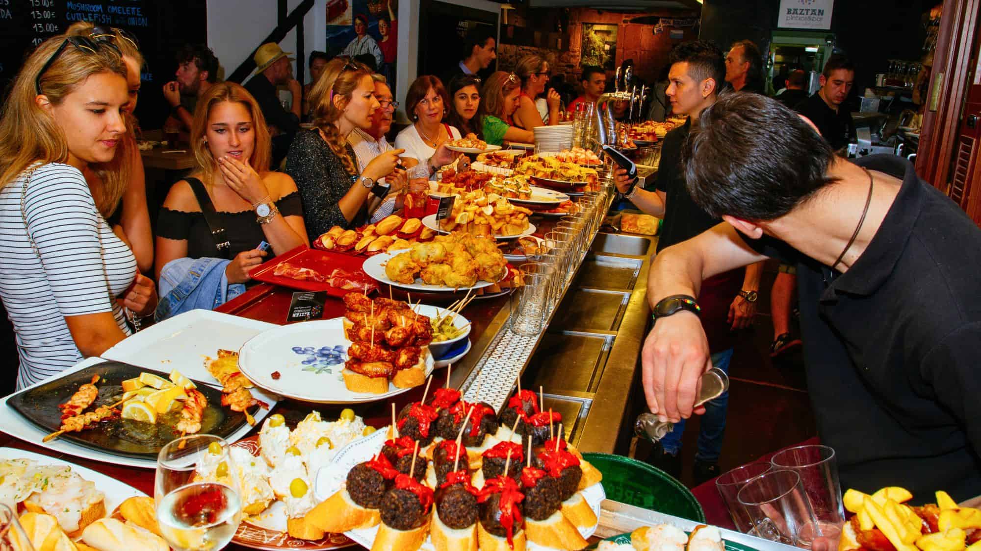 A crowded bar in Spain features trays of colorful pintxos including skewered meats, fried bites, and seafood, with patrons chatting and ordering at the counter.